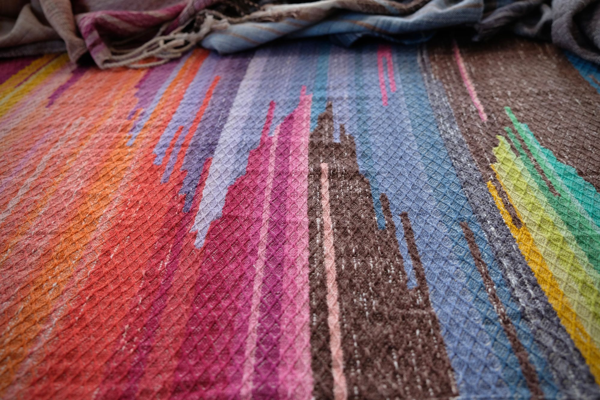 A length of handwoven fabric in earth hues as well as rainbow and sunrise colors lays on a wooden floor