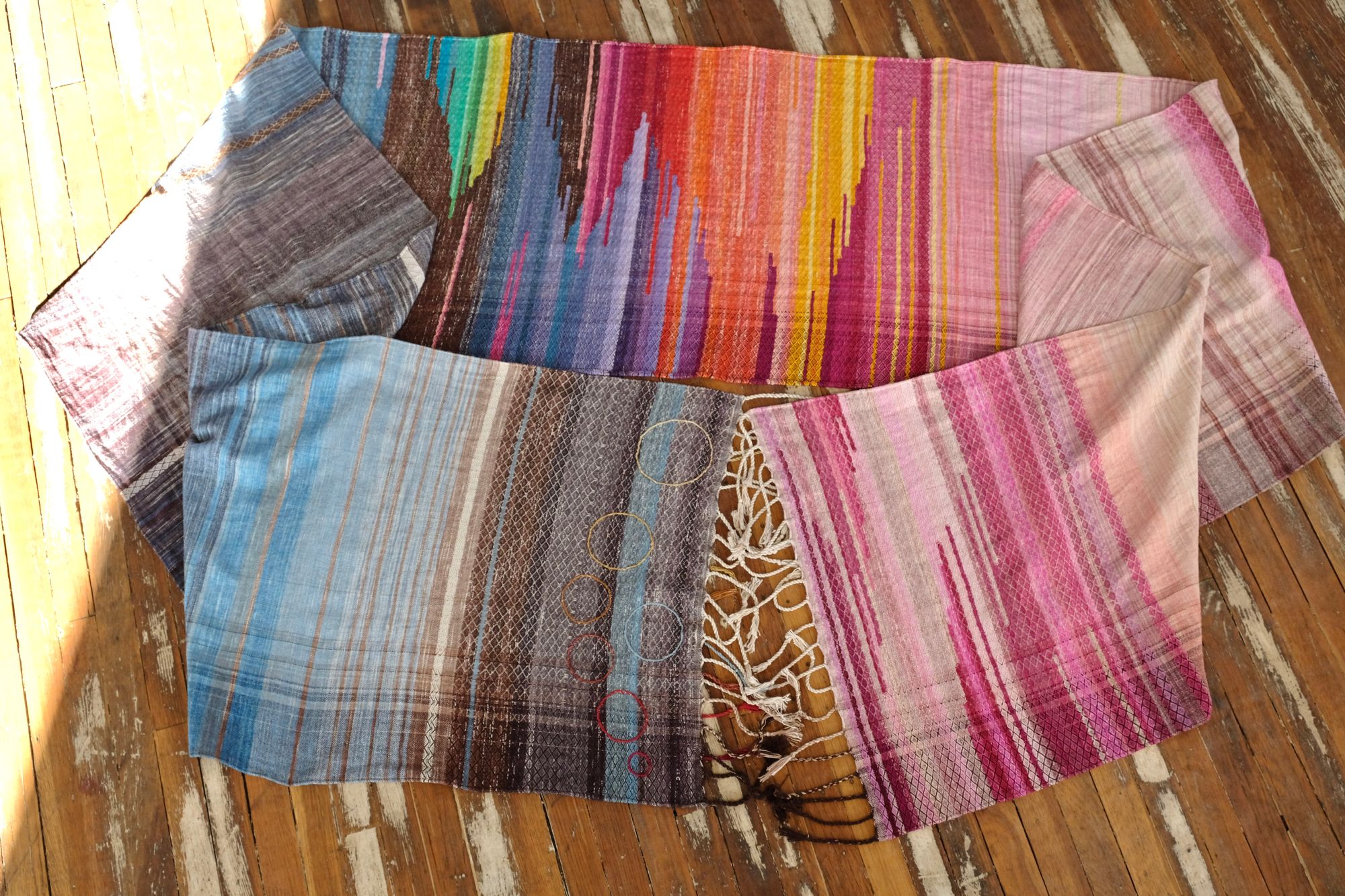 A length of handwoven fabric in earth hues as well as rainbow and sunrise colors lays on a wooden floor