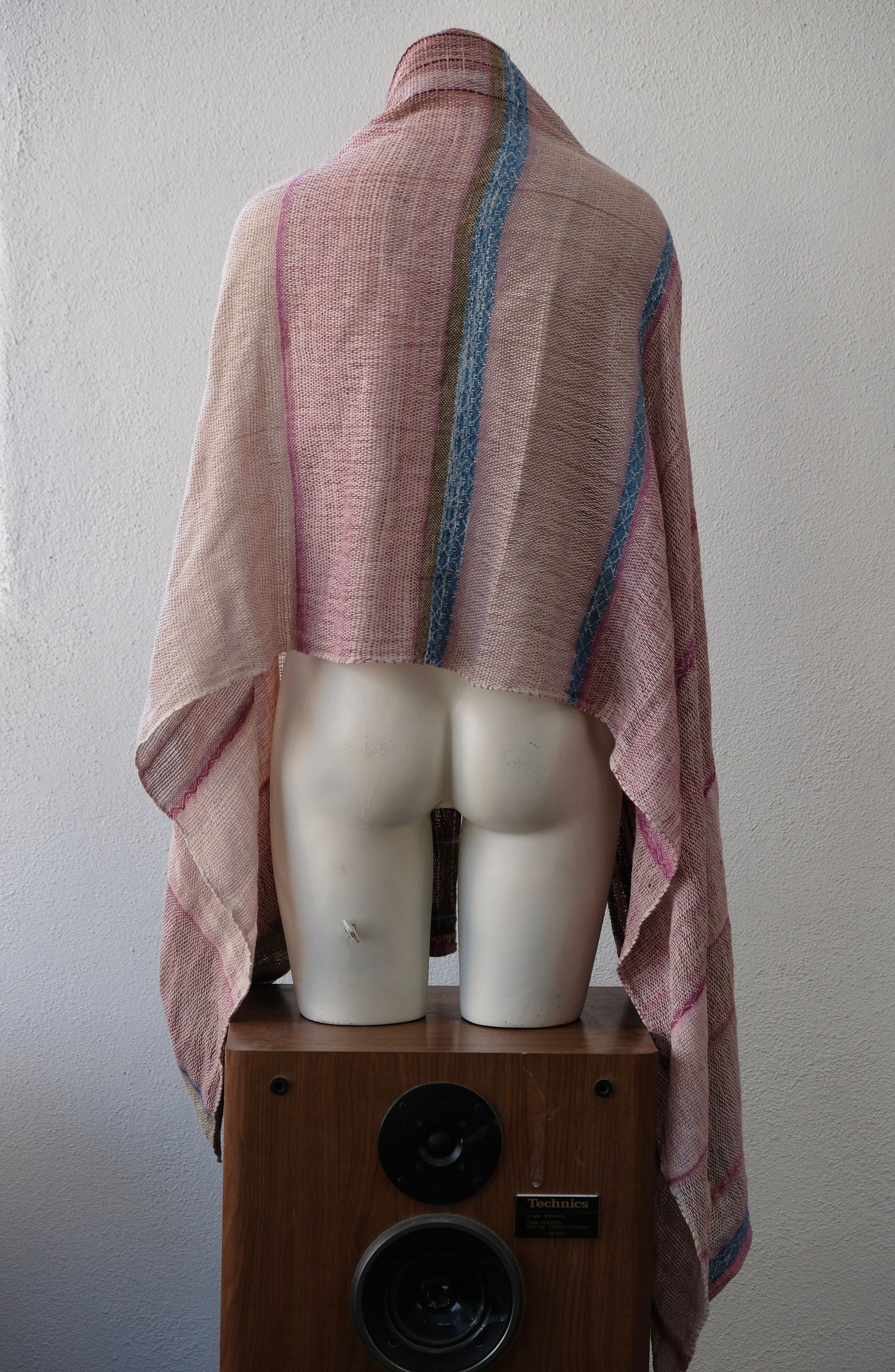 A handwoven scarf shawl loosely woven in naturally dyed earthy pinks and indigo blue rests on a white mannequin which is on a wooden speaker