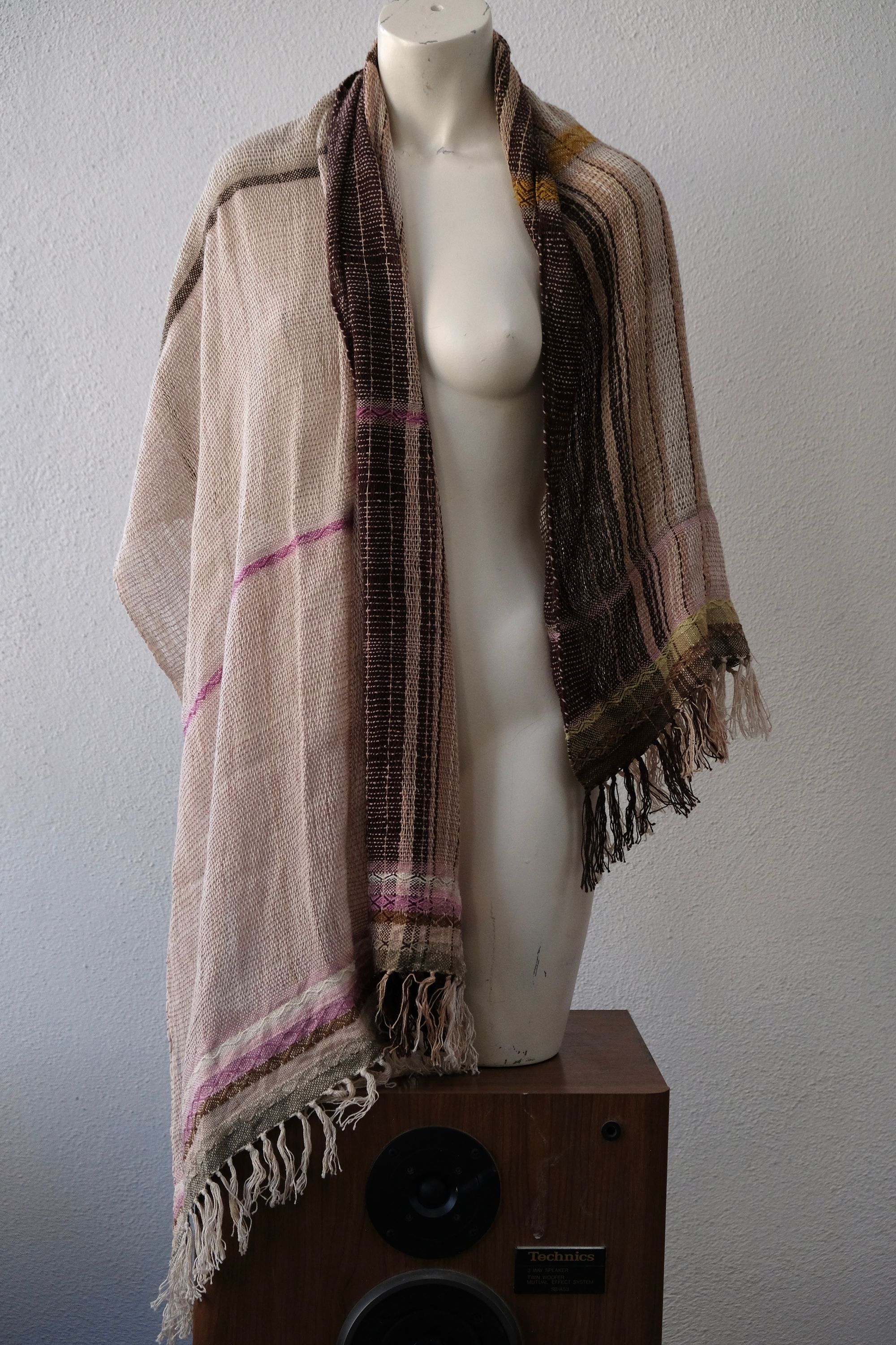 A handwoven scarf shawl loosely woven in naturally dyed earth tones and yellow rests on a white mannequin which is on a wooden speaker