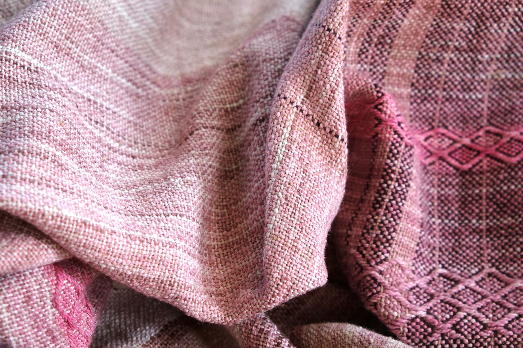 a pink detail of A length of handwoven fabric in earth hues as well as rainbow and sunrise colors lays on a wooden floor