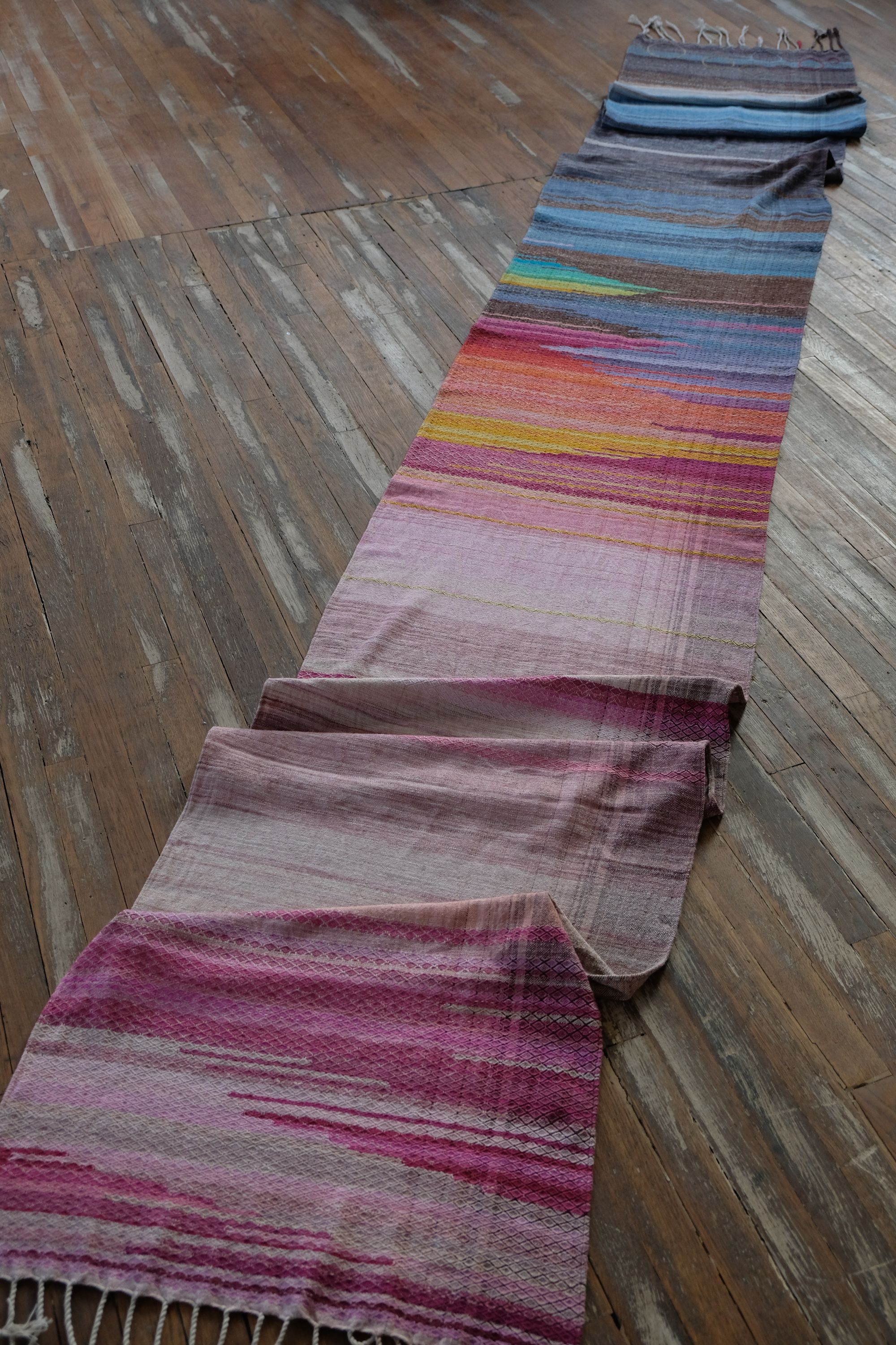 A length of handwoven fabric in earth hues as well as rainbow and sunrise colors lays on a wooden floor