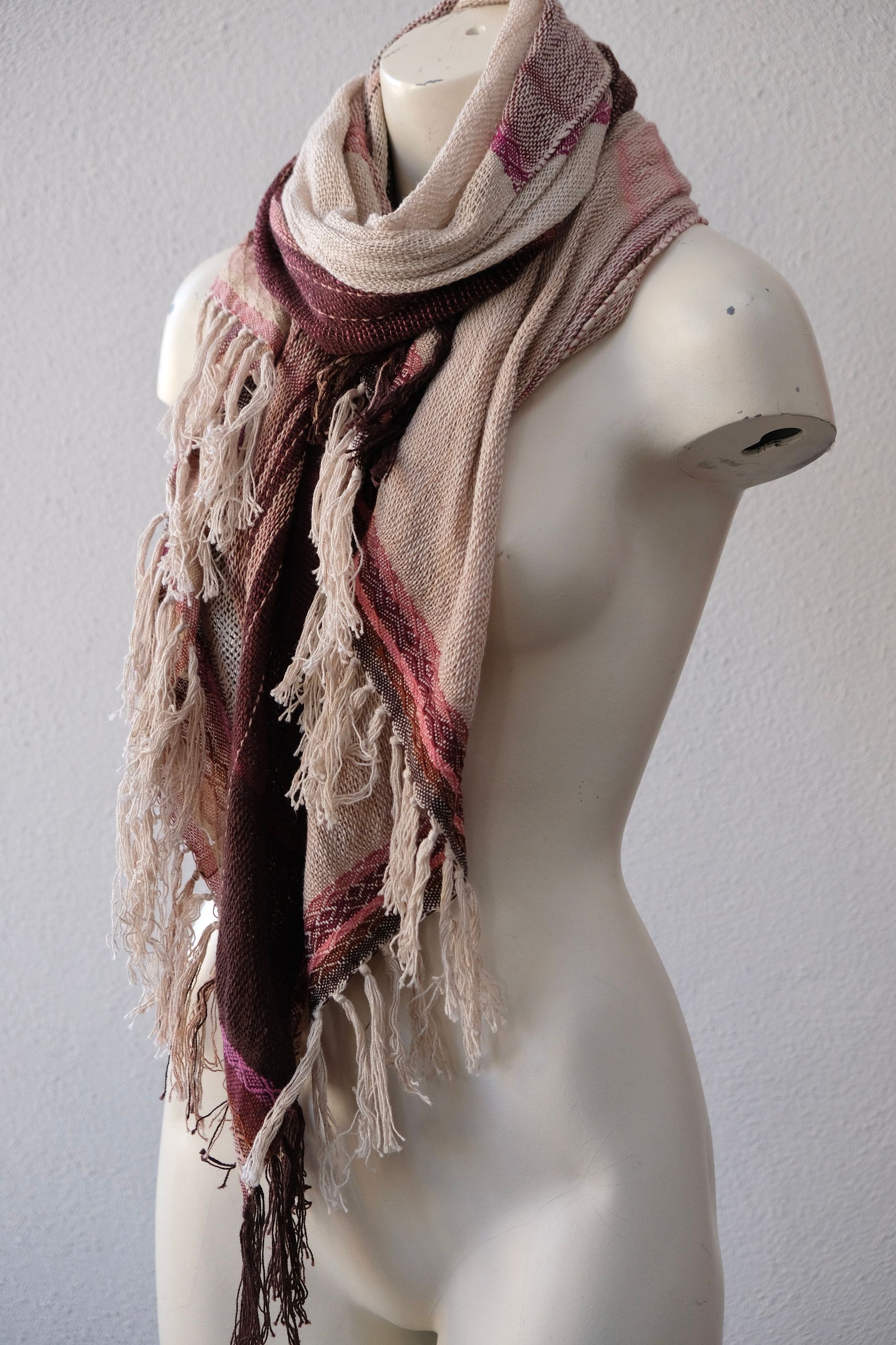 A handwoven scarf shawl loosely woven in a menagerie of creams and pinks rests on a white mannequin which is on a wooden speaker