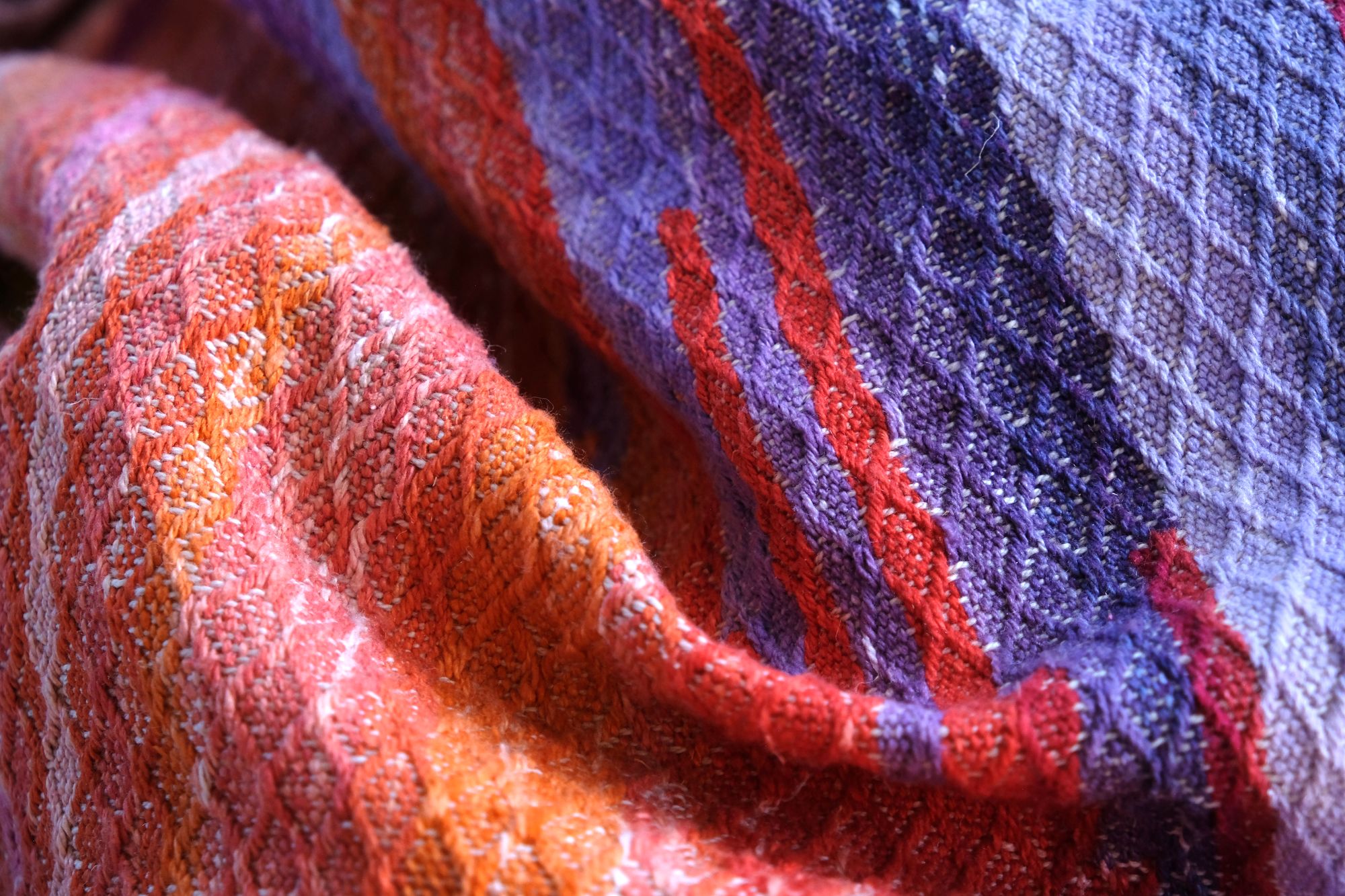 a pink, purple, red and orange tapestry woven detail of A length of handwoven fabric in earth hues as well as rainbow and sunrise colors lays on a wooden floor