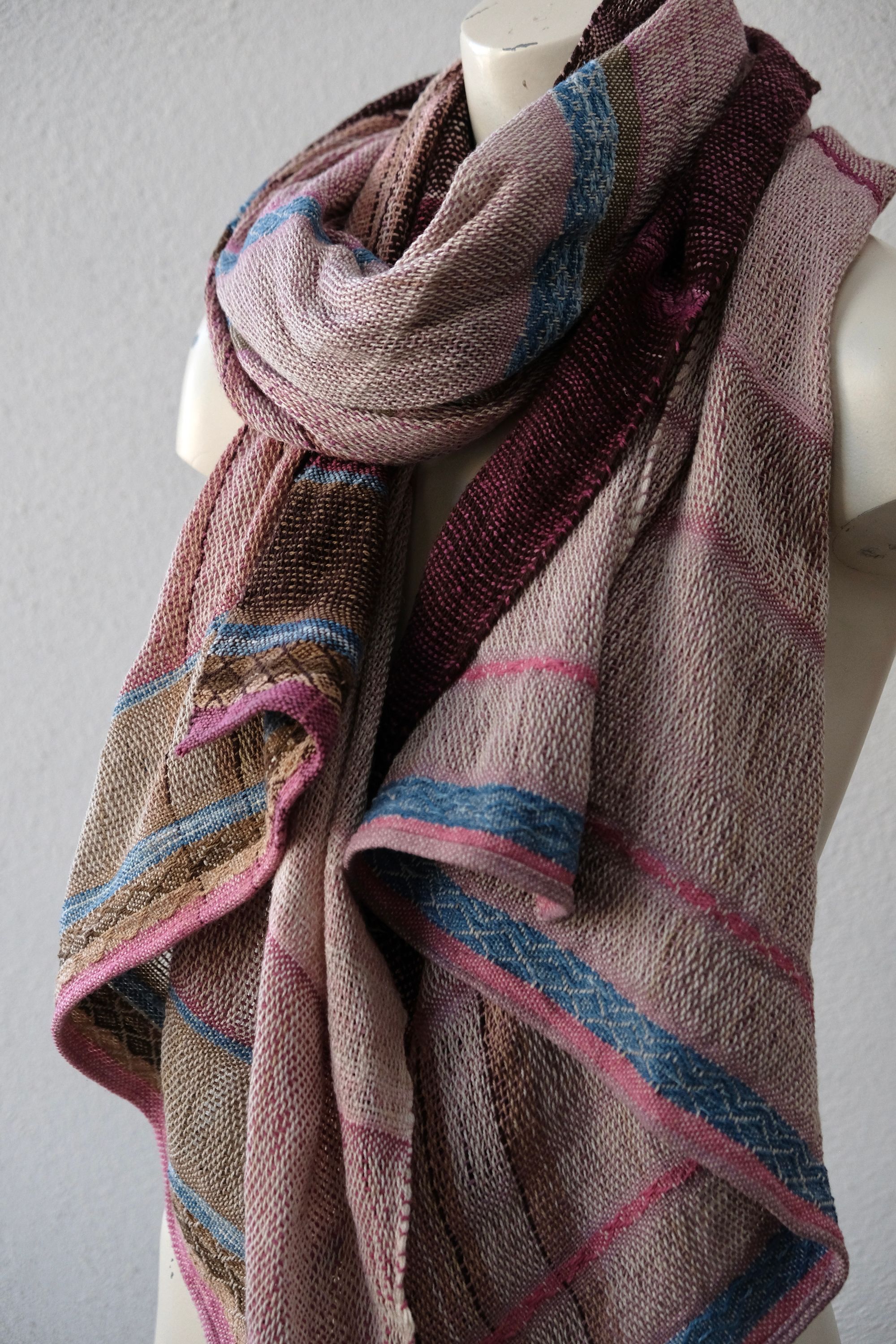 A handwoven scarf shawl loosely woven in naturally dyed earthy pinks and indigo blue rests on a white mannequin which is on a wooden speaker