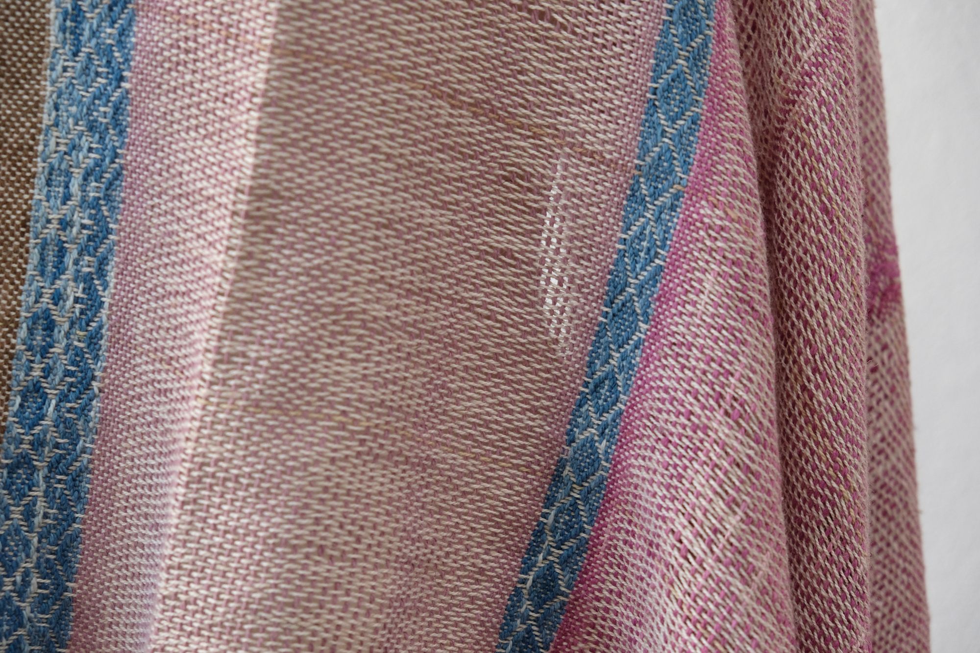 A handwoven scarf shawl loosely woven in naturally dyed earthy pinks and indigo blue rests on a white mannequin which is on a wooden speaker