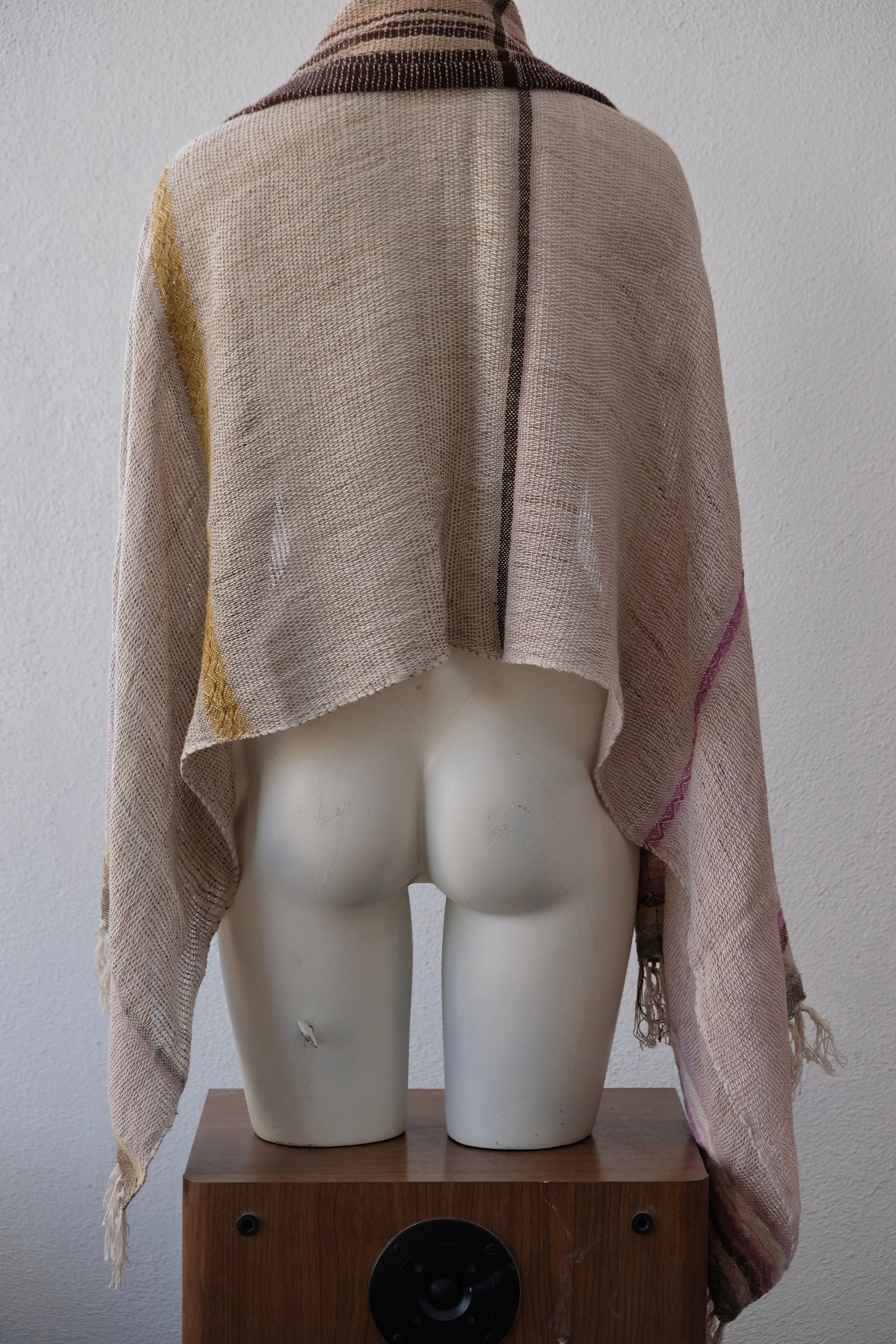 A handwoven scarf shawl loosely woven in naturally dyed earth tones and yellow rests on a white mannequin which is on a wooden speaker