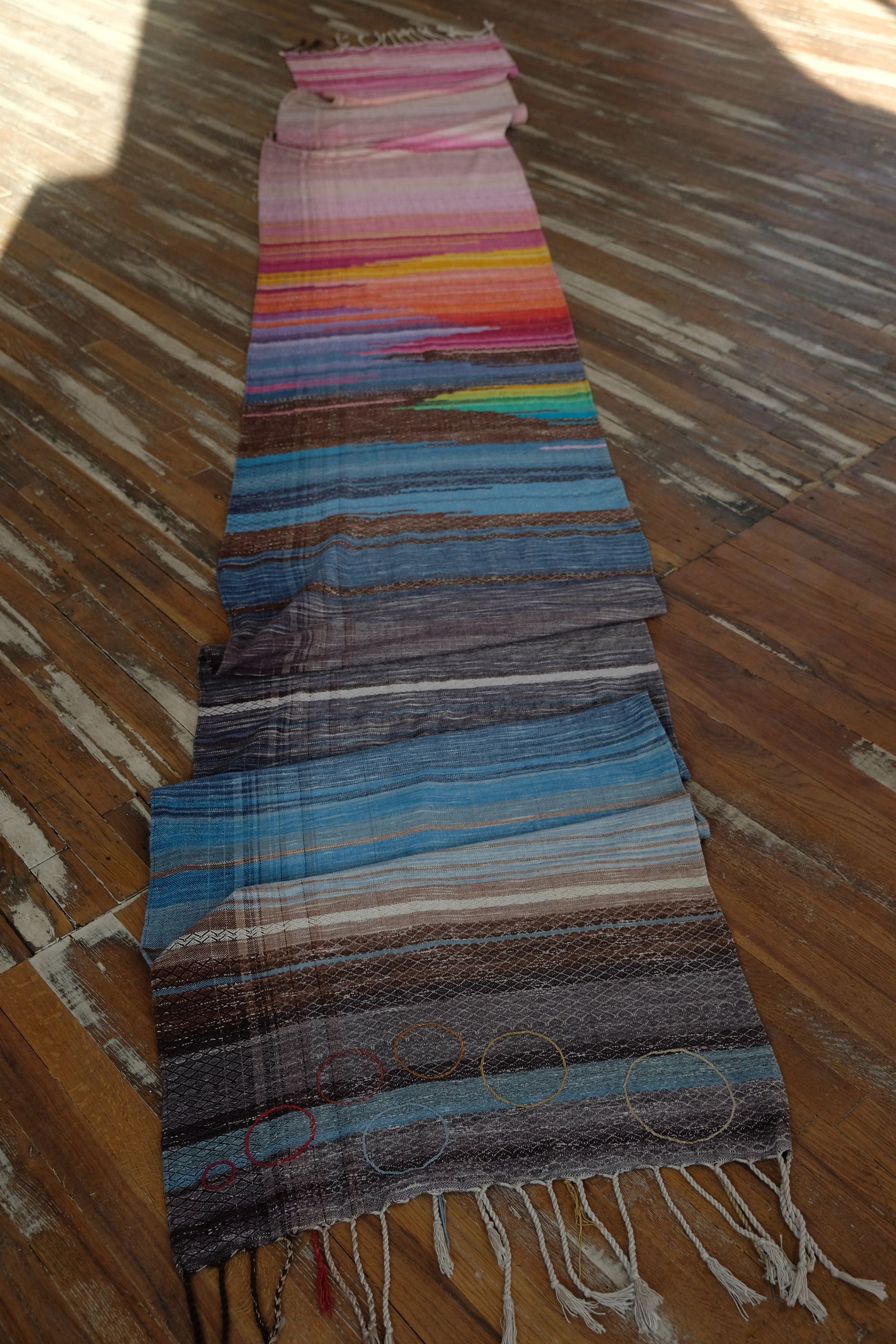 A length of handwoven fabric in earth hues as well as rainbow and sunrise colors lays on a wooden floor