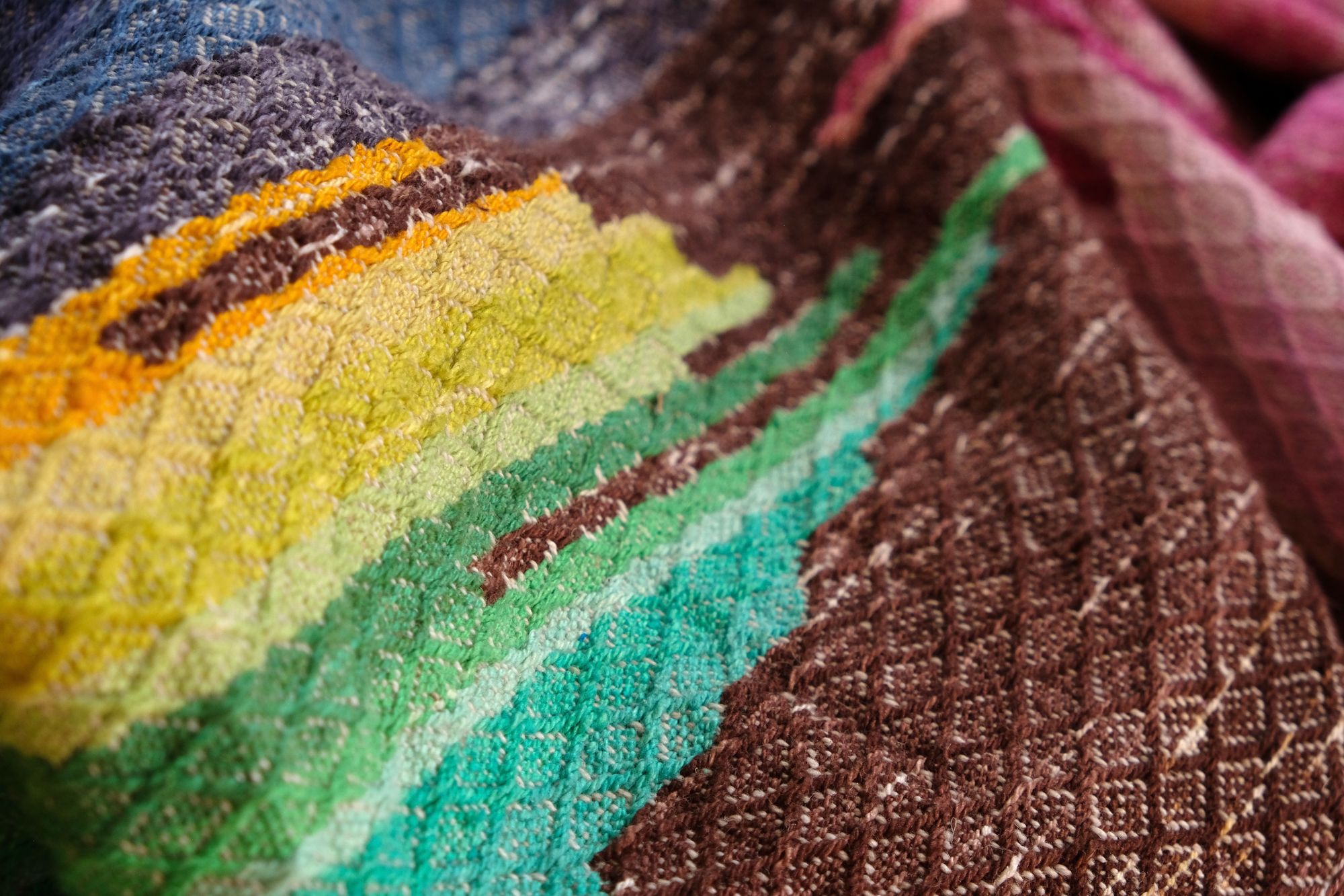 a green, blue, yellow and brown tapestry woven detail of A length of handwoven fabric in earth hues as well as rainbow and sunrise colors lays on a wooden floor