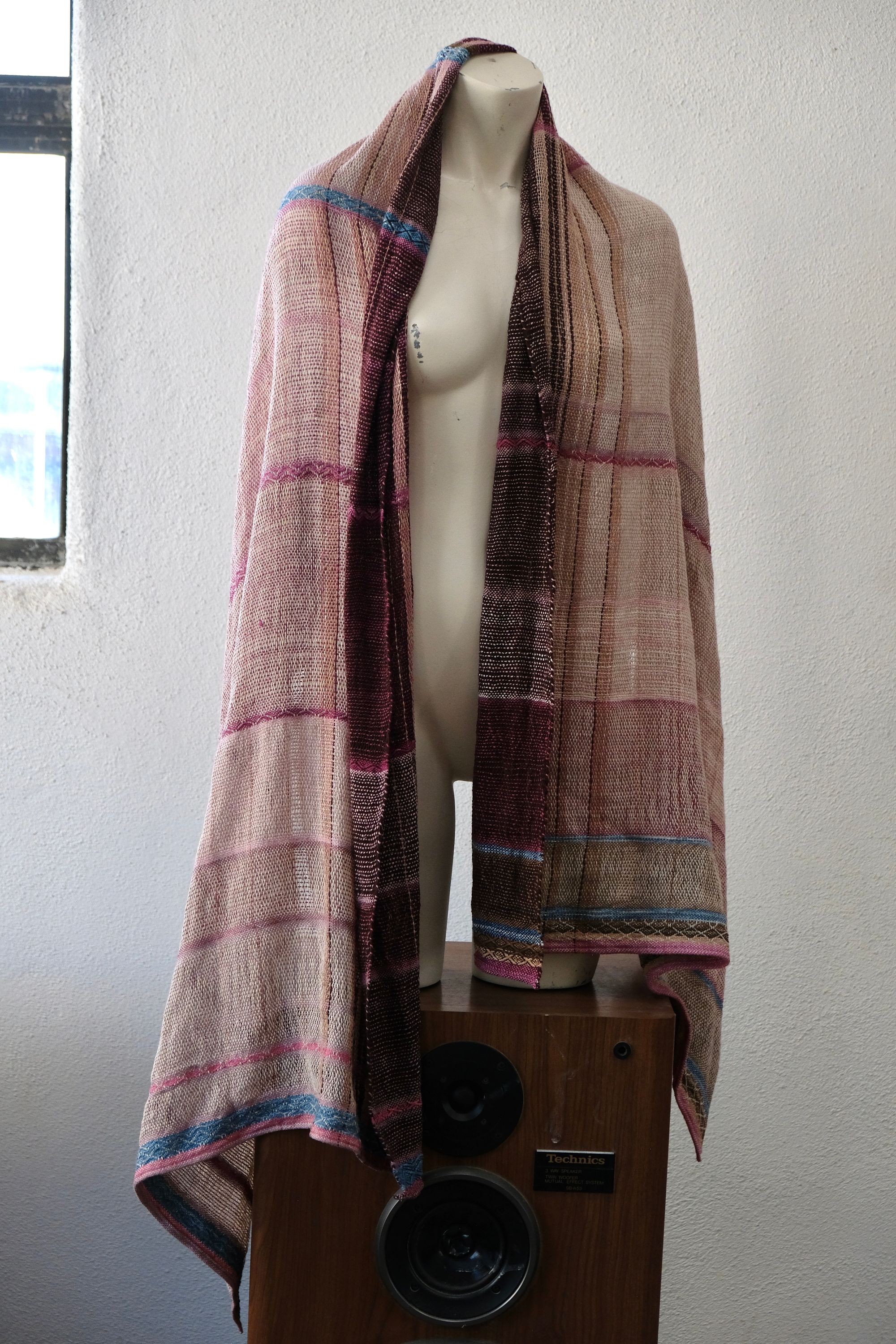 A handwoven scarf shawl loosely woven in naturally dyed earthy pinks and indigo blue rests on a white mannequin which is on a wooden speaker