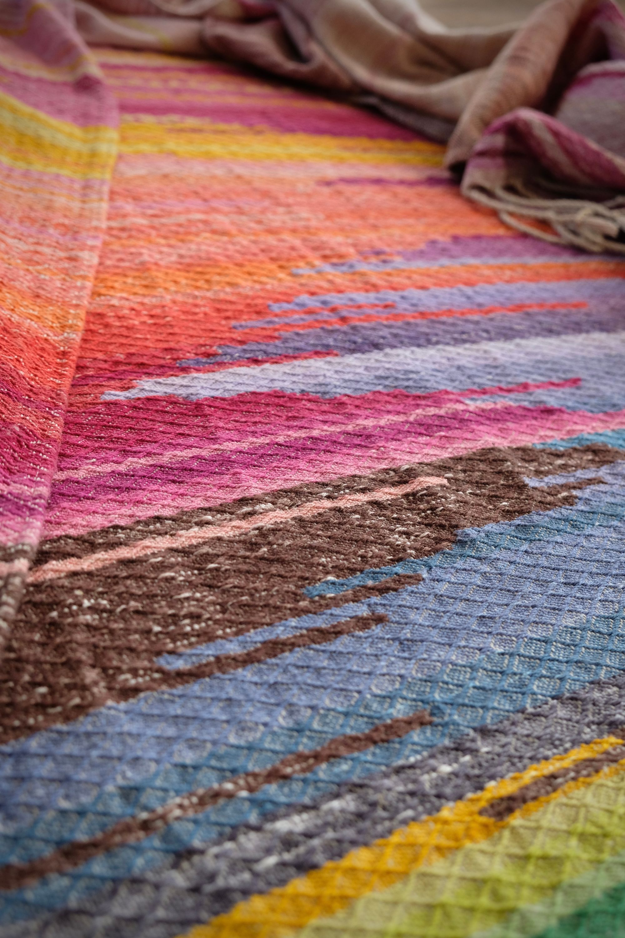 A length of handwoven fabric in earth hues as well as rainbow and sunrise colors lays on a wooden floor