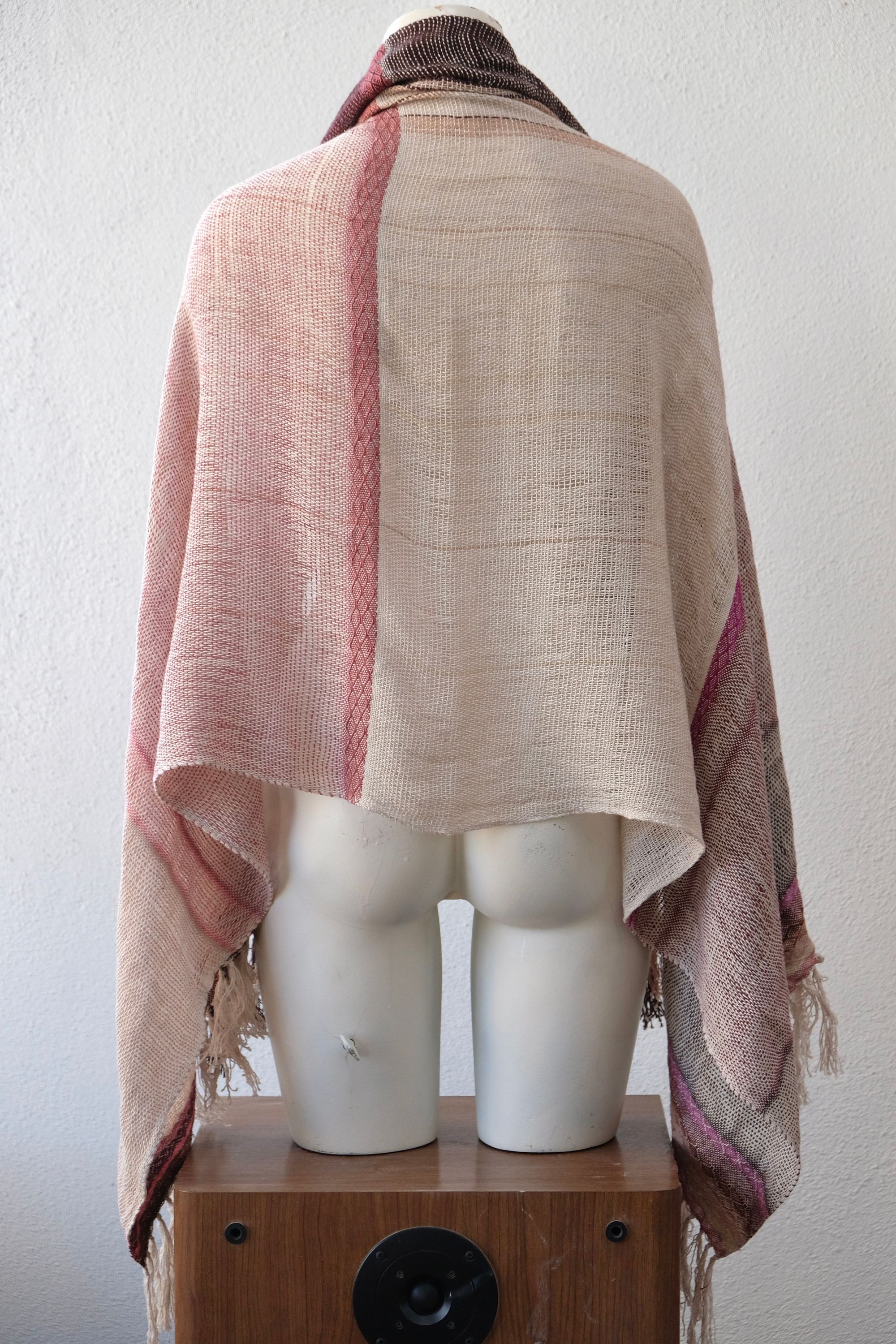 A handwoven scarf shawl loosely woven in a menagerie of creams and pinks rests on a white mannequin which is on a wooden speaker