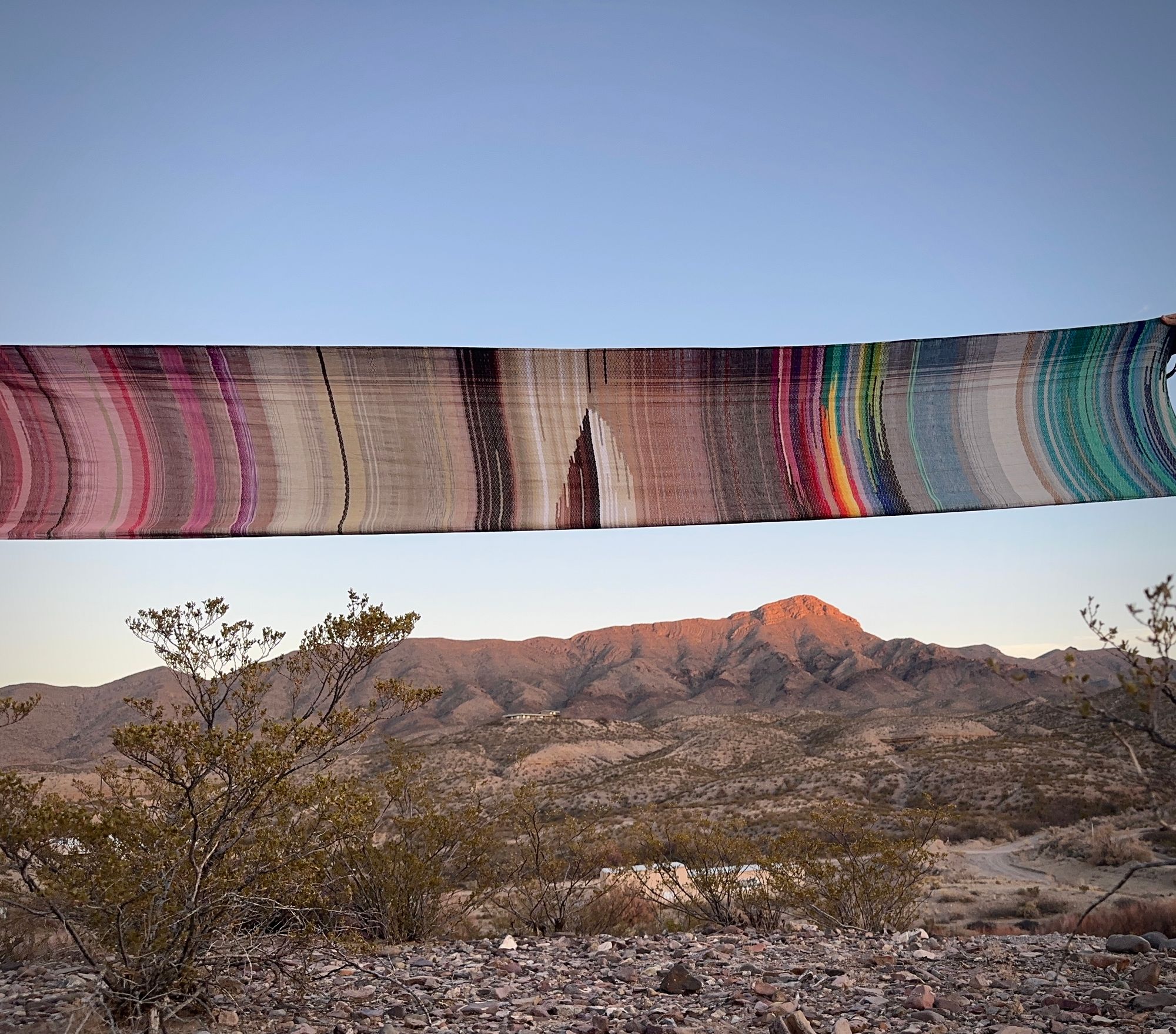 5 m of head woven rainbow colored fabric is pulled across the sky above a horizon of a desert mountain, bathed in golden sunset light