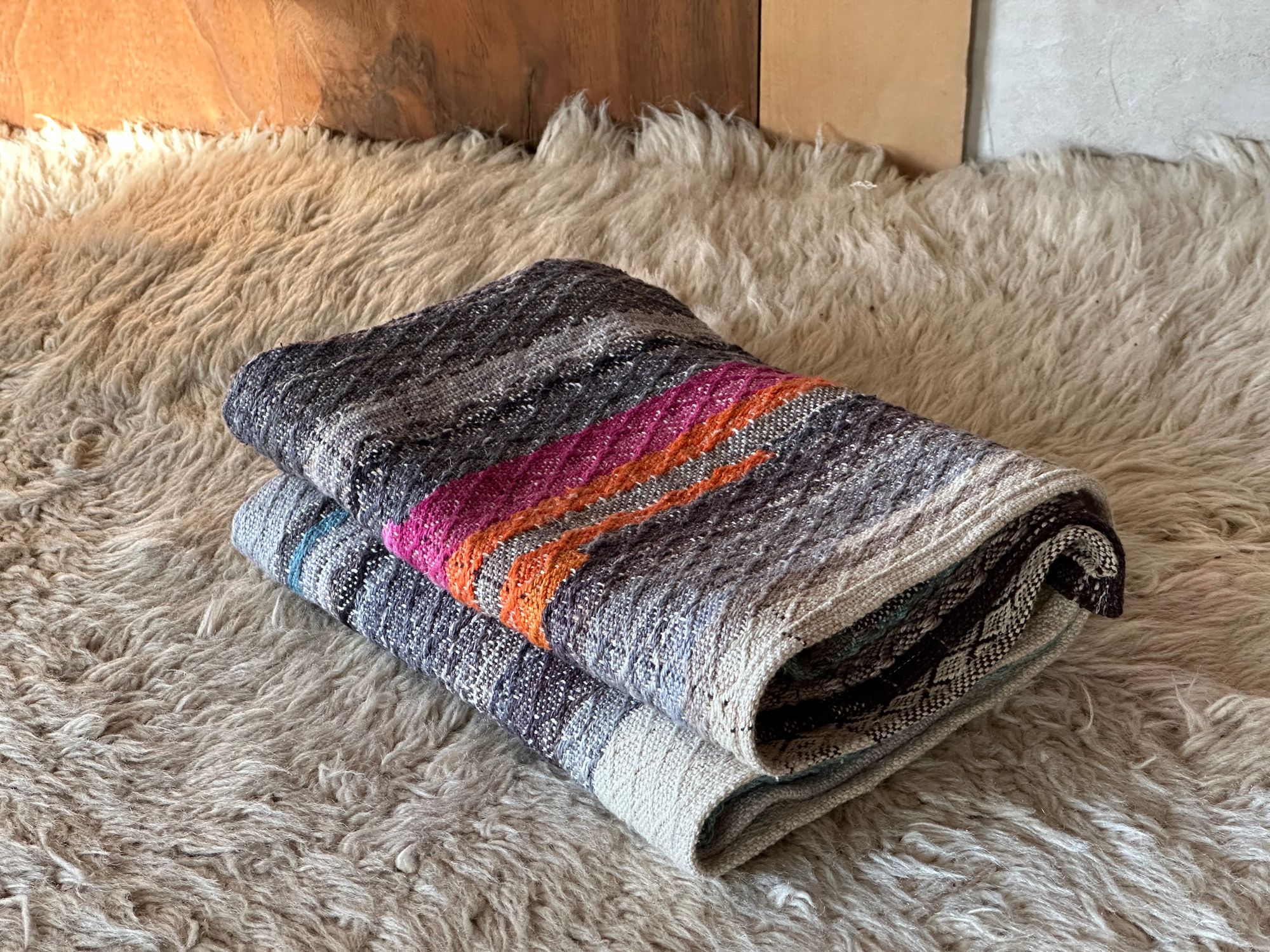 A small handwoven blanket with a diamond pattern in blues and grey laying on a white furry rug 