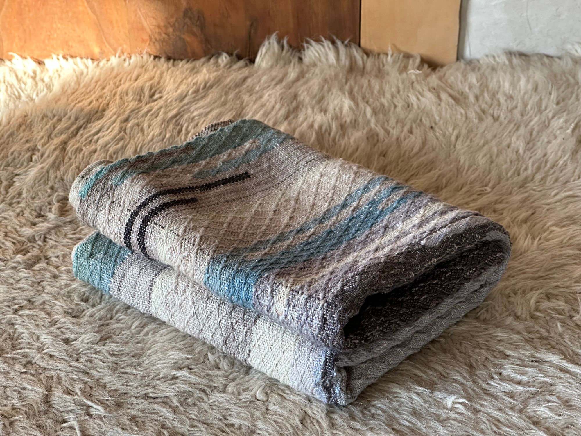 A small handwoven blanket with a diamond pattern in blues and grey laying on a white furry rug 