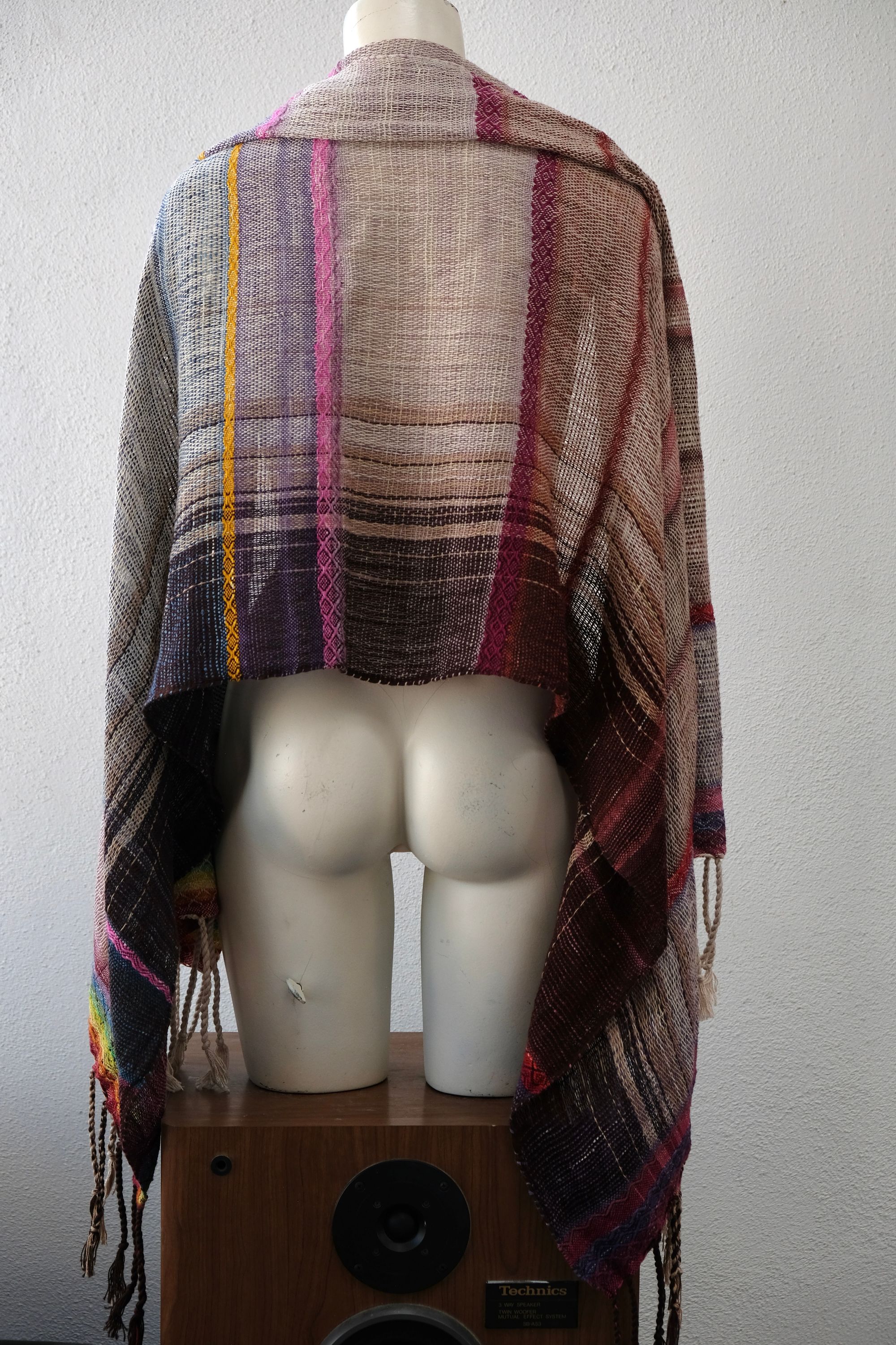 A white painted mannequin stands on a wooden speaker wearing an etherial handwoven scarf with a mixture of loose weave and rainbow shades, in rainbow hues