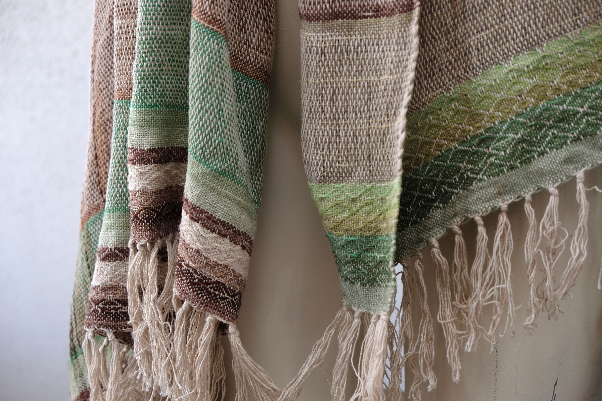 A white painted mannequin stands on a wooden speaker wearing an etherial handwoven scarf in greens and browns