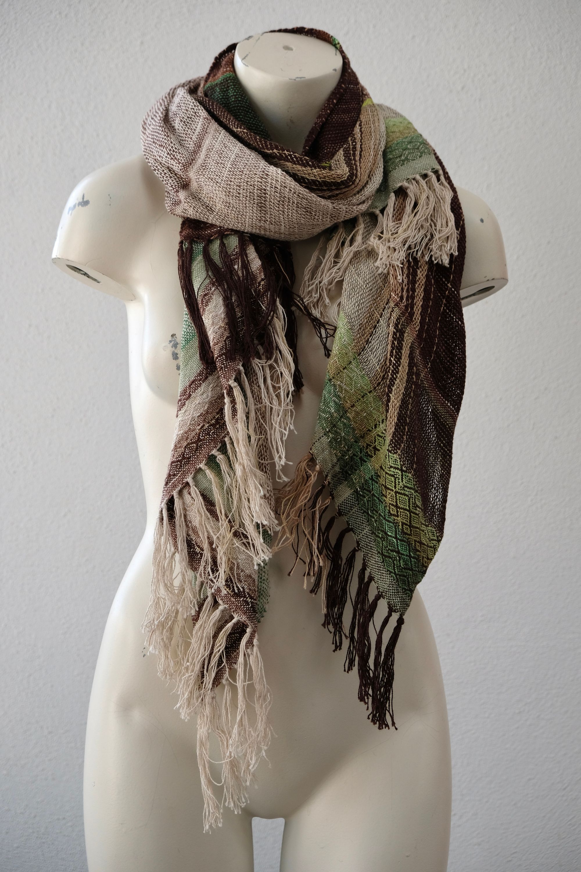 A white painted mannequin stands on a wooden speaker wearing an etherial handwoven scarf in greens and browns