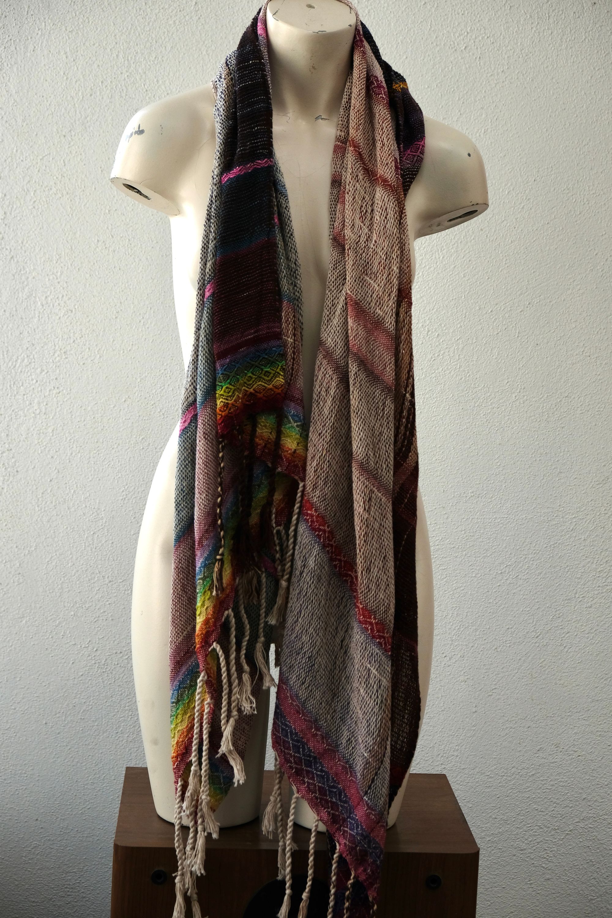 A white painted mannequin stands on a wooden speaker wearing an etherial handwoven scarf with a mixture of loose weave and rainbow shades, in rainbow hues