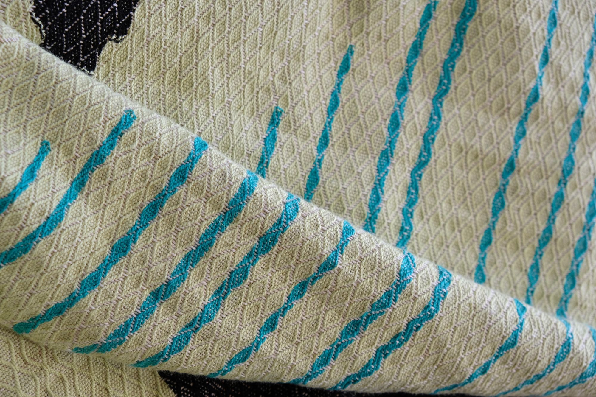 Handwoven diamond pattern fabric in shades of mint green, cream, black, turquoise with stripes of rainbow lays on a wooden floor.