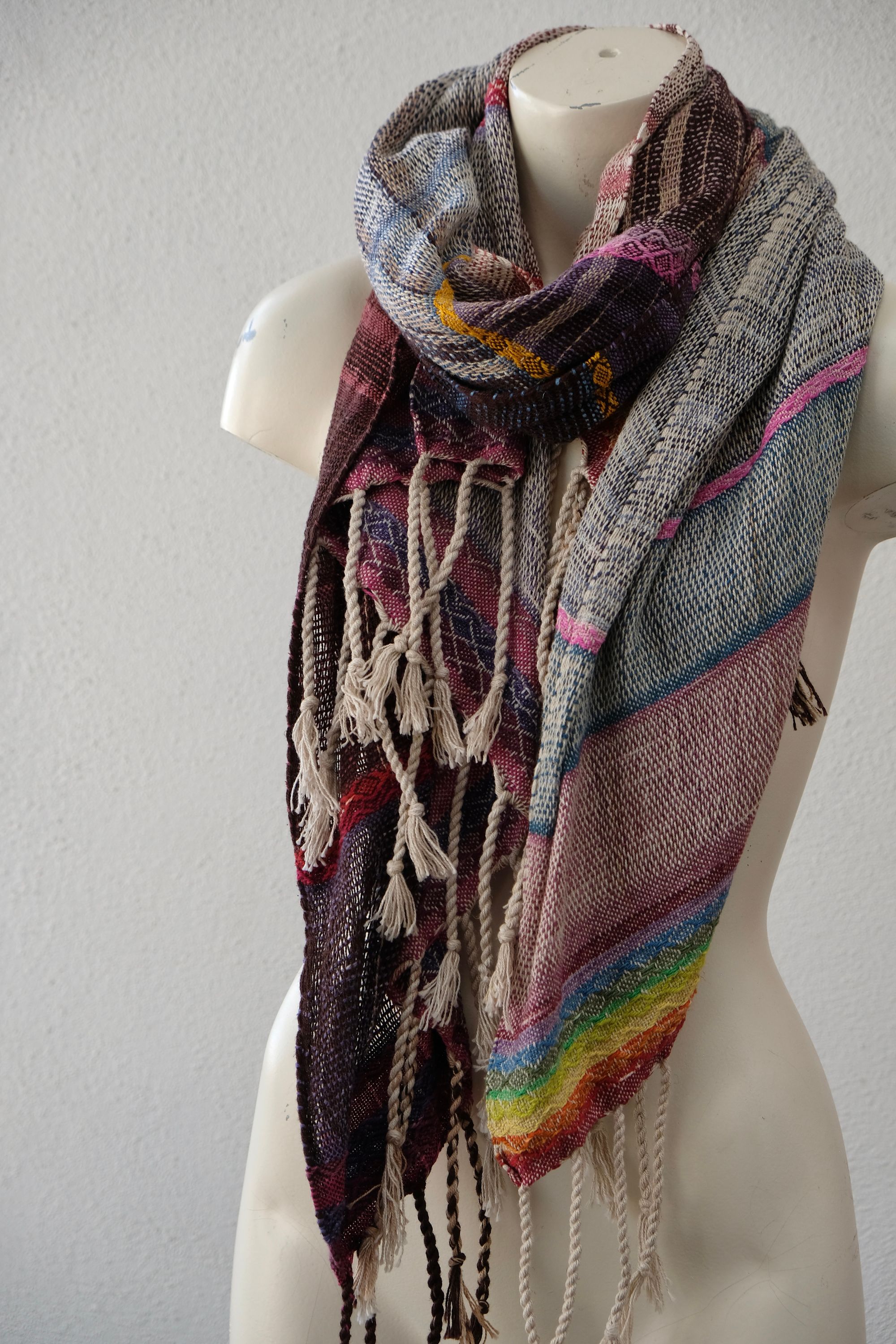 A white painted mannequin stands on a wooden speaker wearing an etherial handwoven scarf with a mixture of loose weave and rainbow shades, in rainbow hues