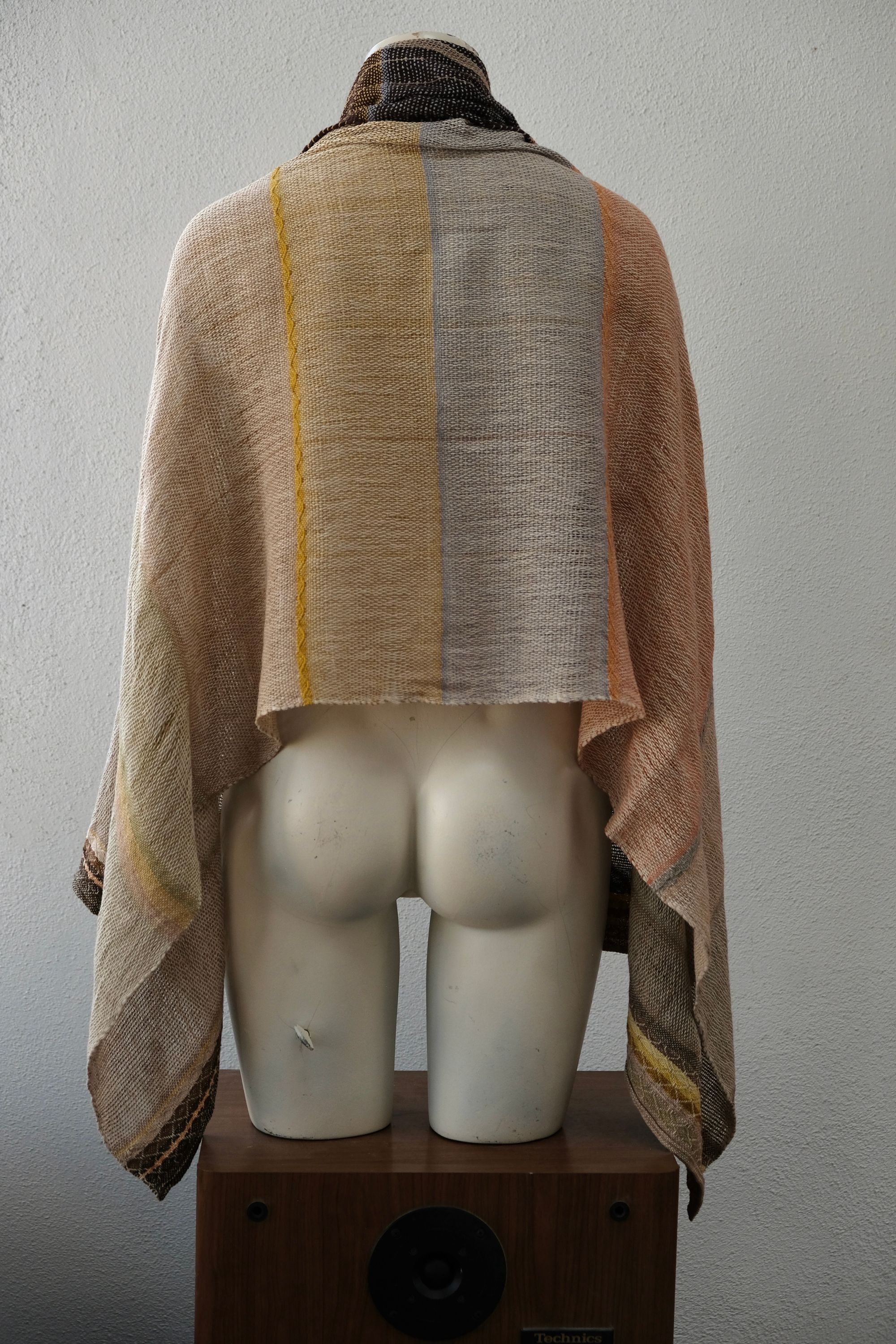 A white painted mannequin stands on a wooden speaker wearing an etherial handwoven scarf with a mixture of loose weave and naturally dyed bright earthy shades of yellow, brown, blue, orange, peach and green