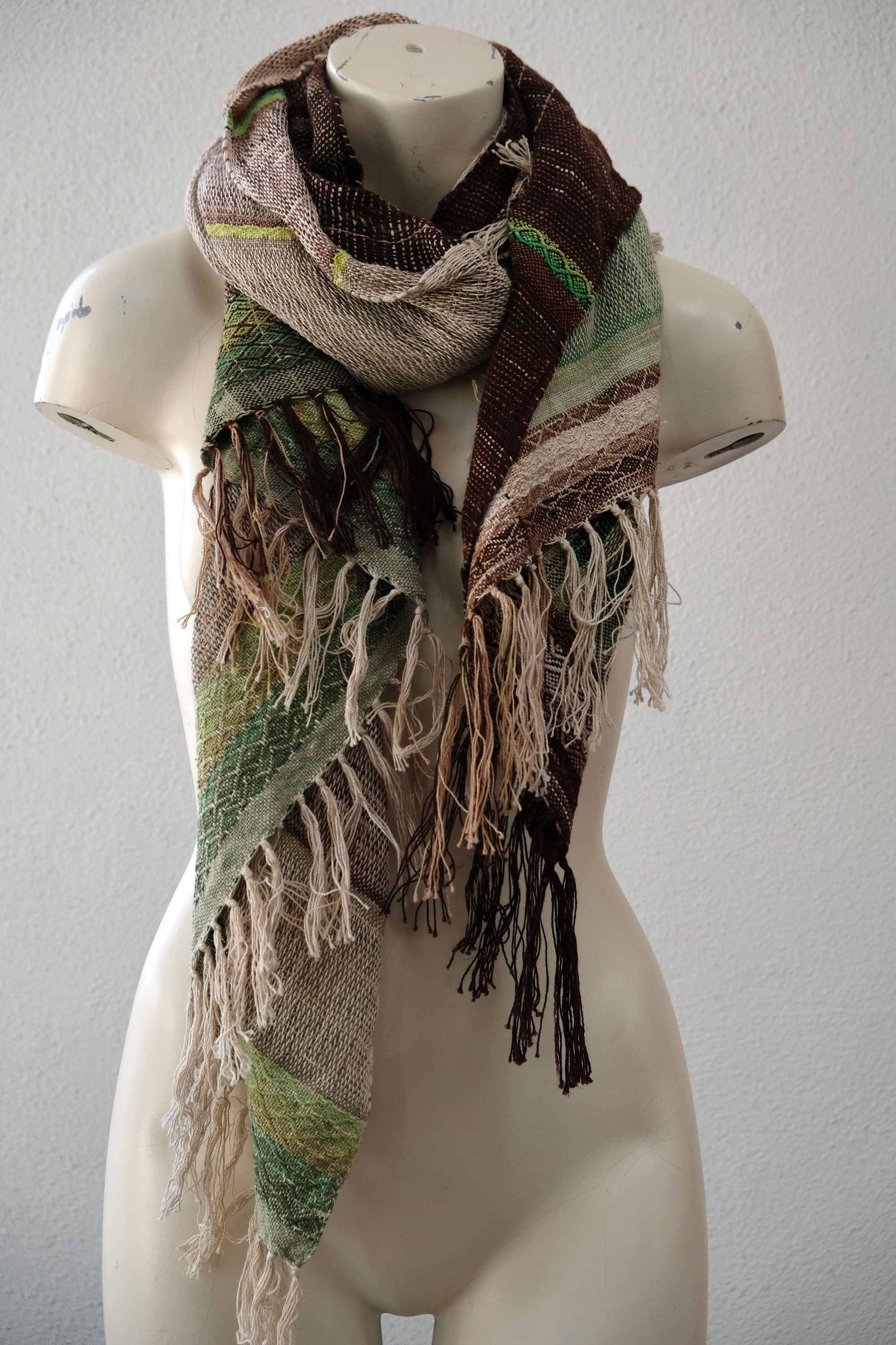 A white painted mannequin stands on a wooden speaker wearing an etherial handwoven scarf in greens and browns