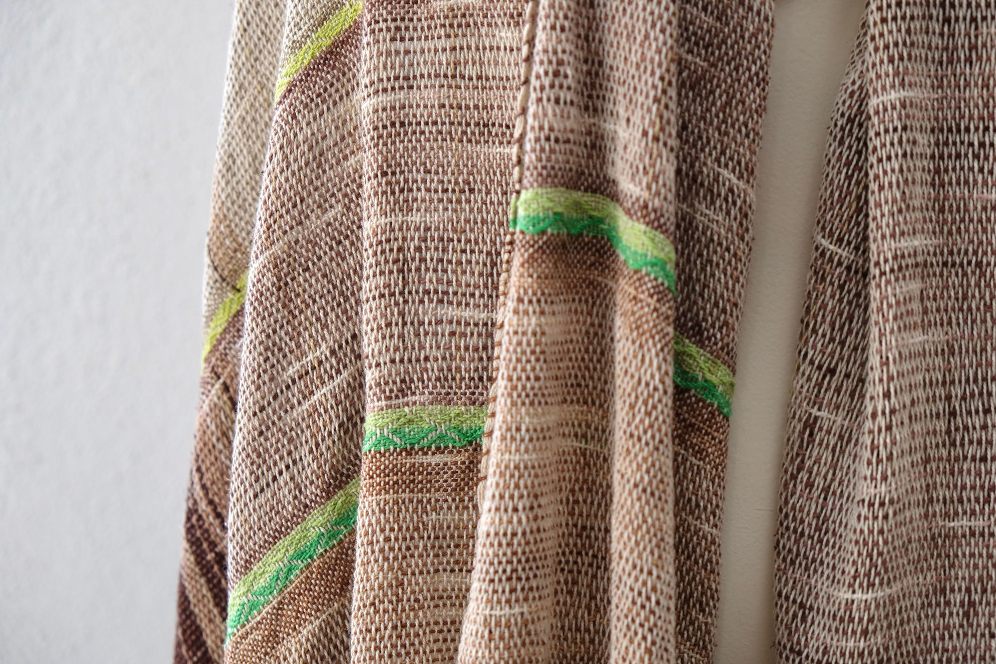 A white painted mannequin stands on a wooden speaker wearing an etherial handwoven scarf in greens and browns