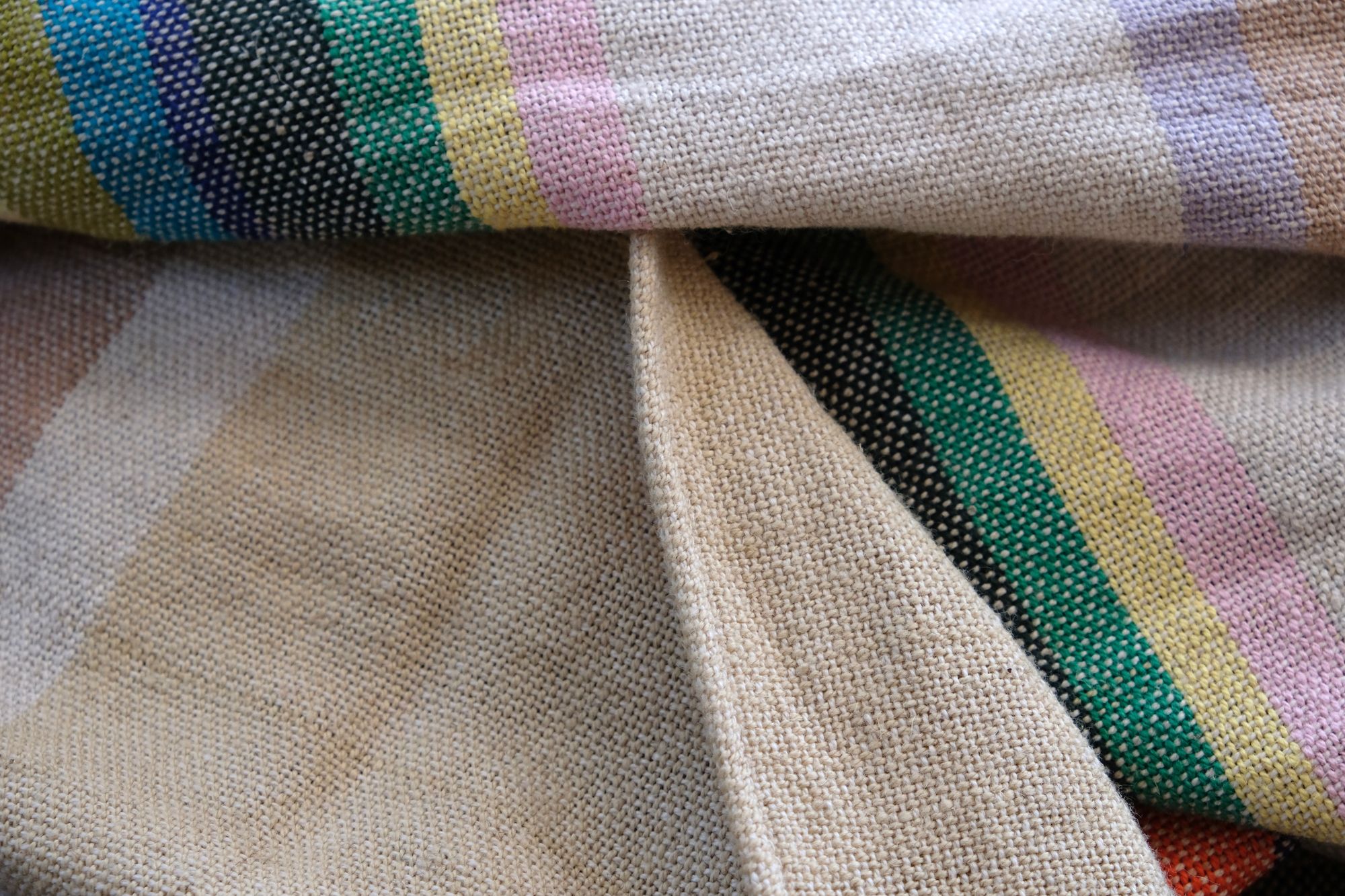 Handwoven diamond pattern fabric in shades of mint green, cream, black, turquoise with stripes of rainbow lays on a wooden floor.