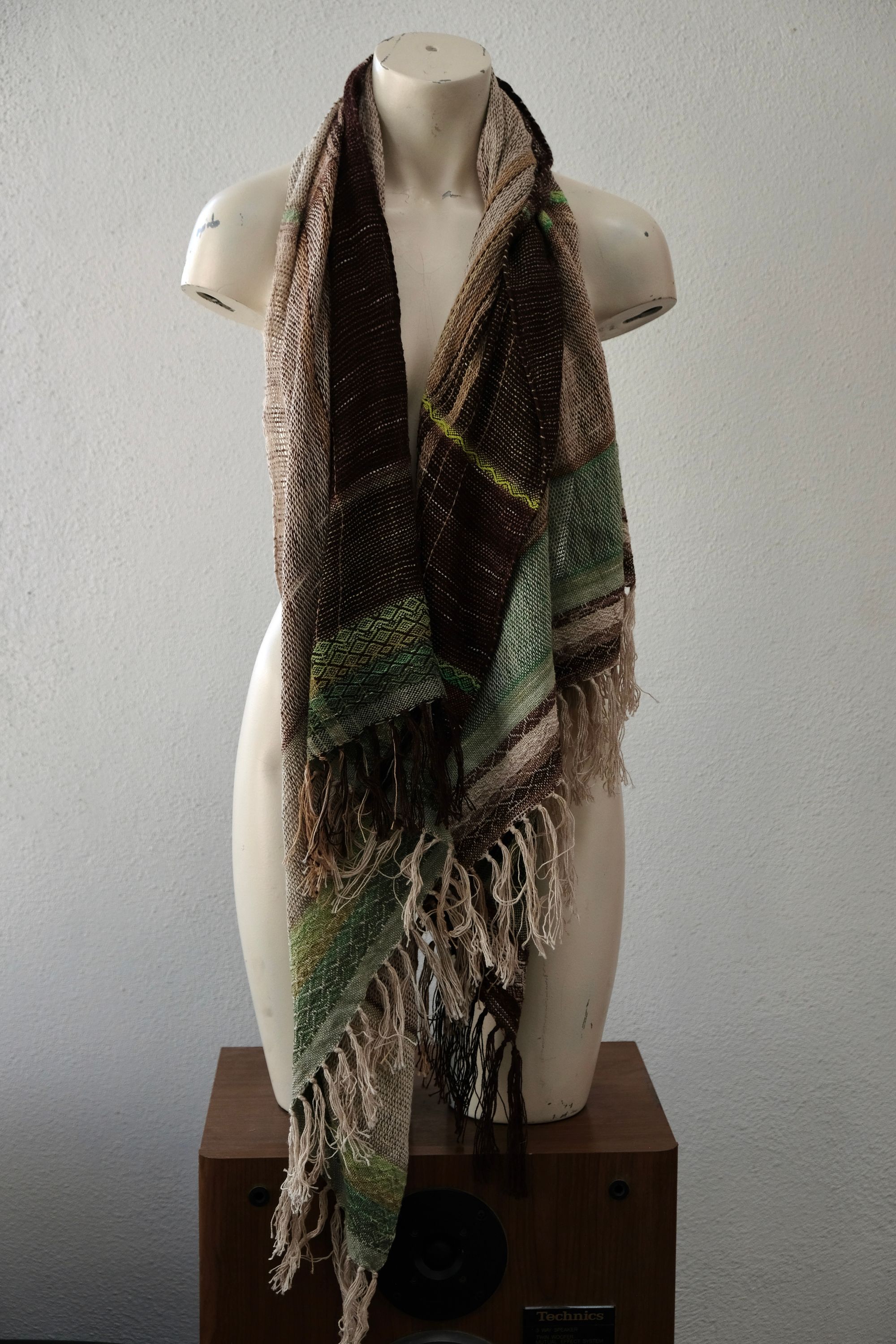 A white painted mannequin stands on a wooden speaker wearing an etherial handwoven scarf in greens and browns