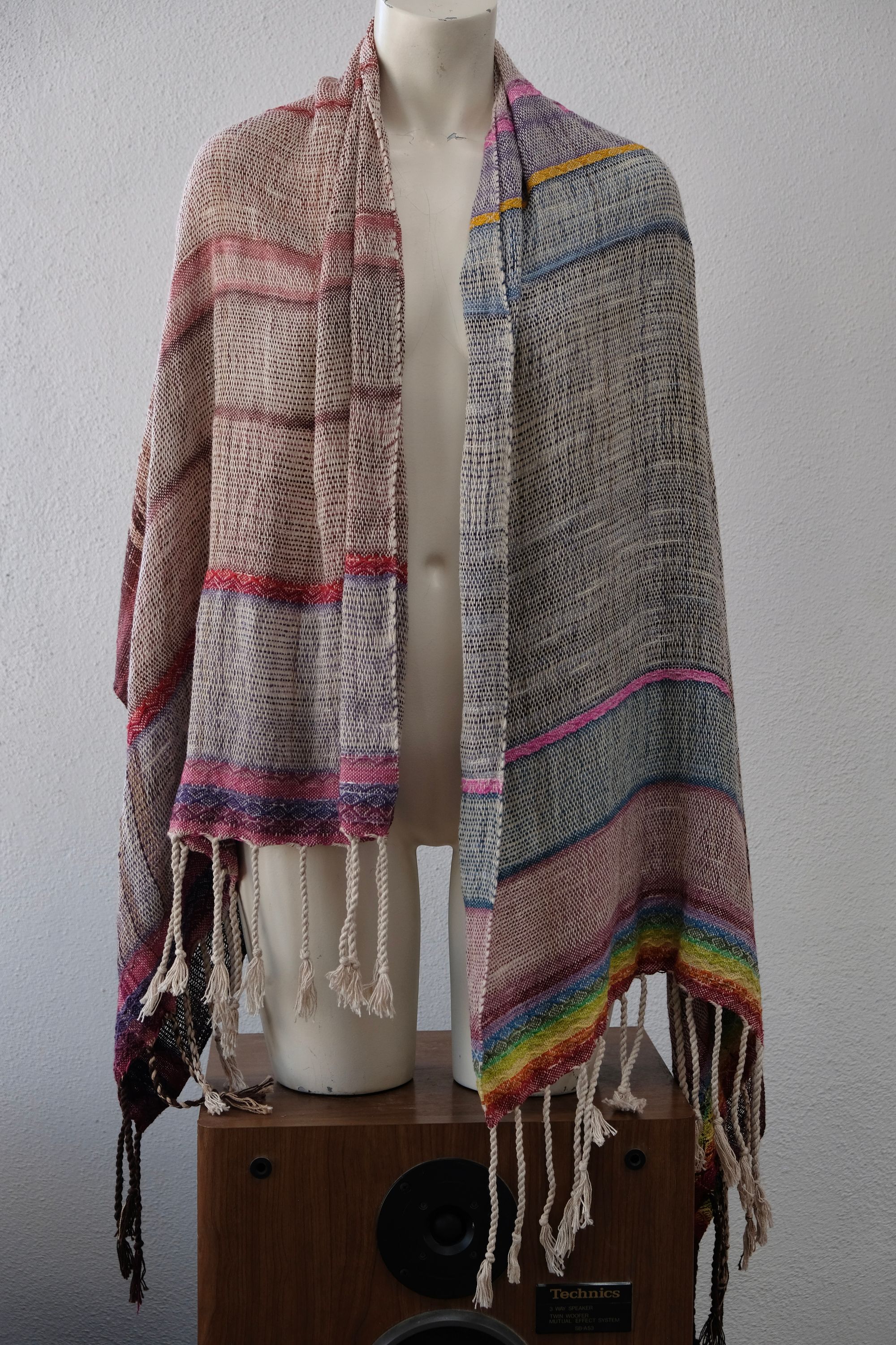 A white painted mannequin stands on a wooden speaker wearing an etherial handwoven scarf with a mixture of loose weave and rainbow shades, in rainbow hues