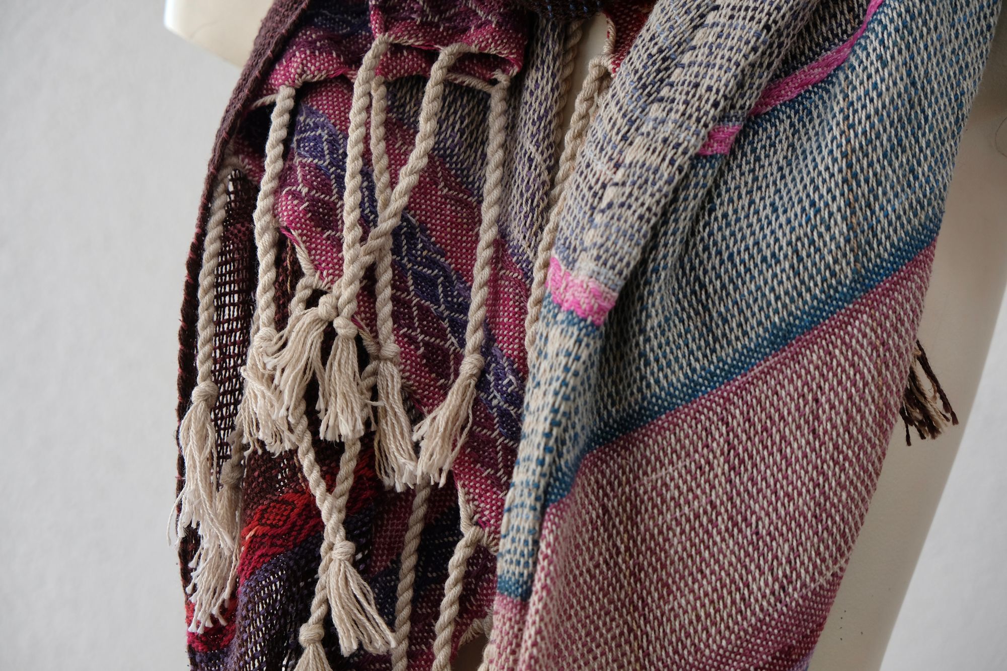 A white painted mannequin stands on a wooden speaker wearing an etherial handwoven scarf with a mixture of loose weave and rainbow shades, in rainbow hues