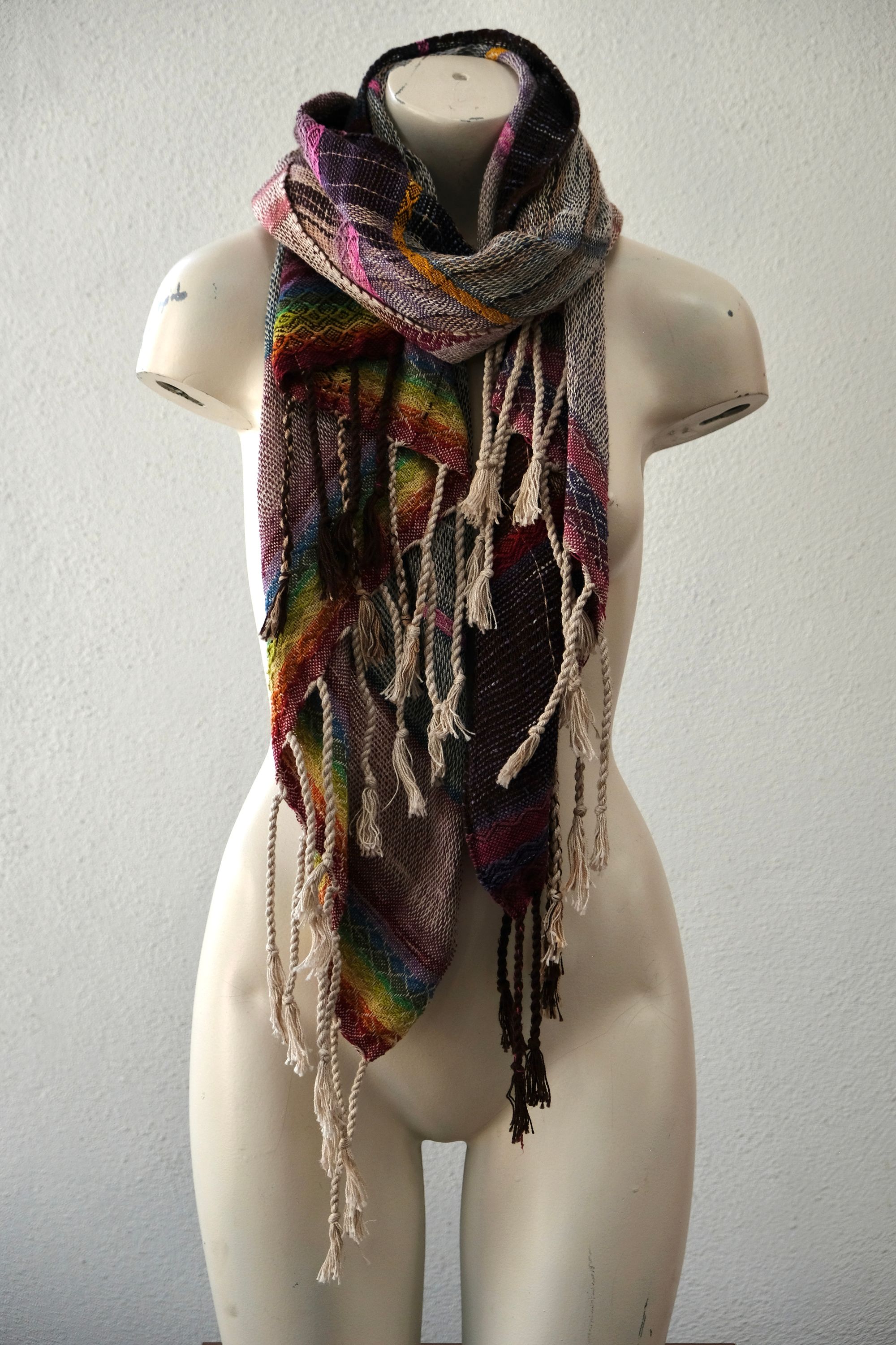 A white painted mannequin stands on a wooden speaker wearing an etherial handwoven scarf with a mixture of loose weave and rainbow shades, in rainbow hues