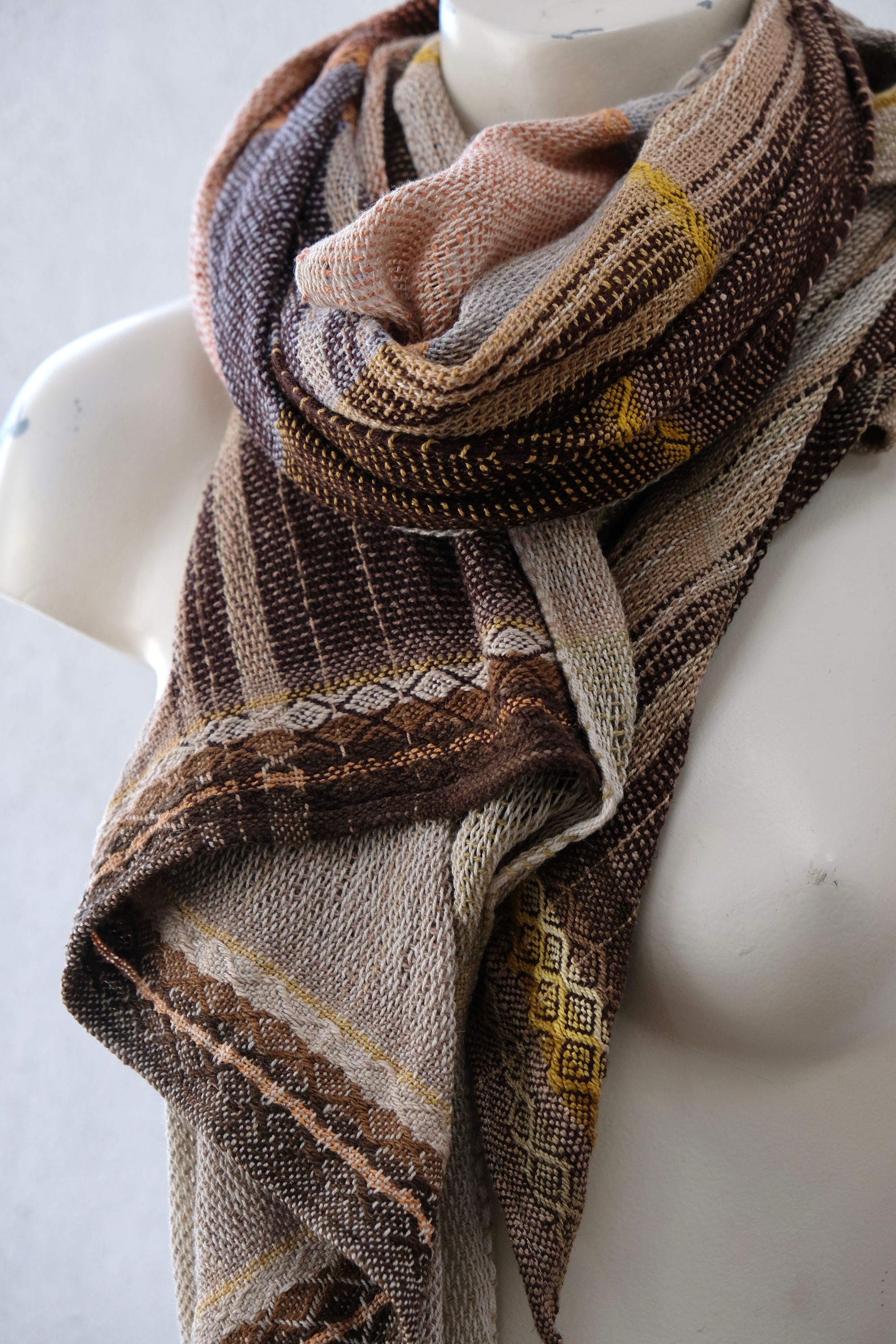 A white painted mannequin stands on a wooden speaker wearing an etherial handwoven scarf with a mixture of loose weave and naturally dyed bright earthy shades of yellow, brown, blue, orange, peach and green