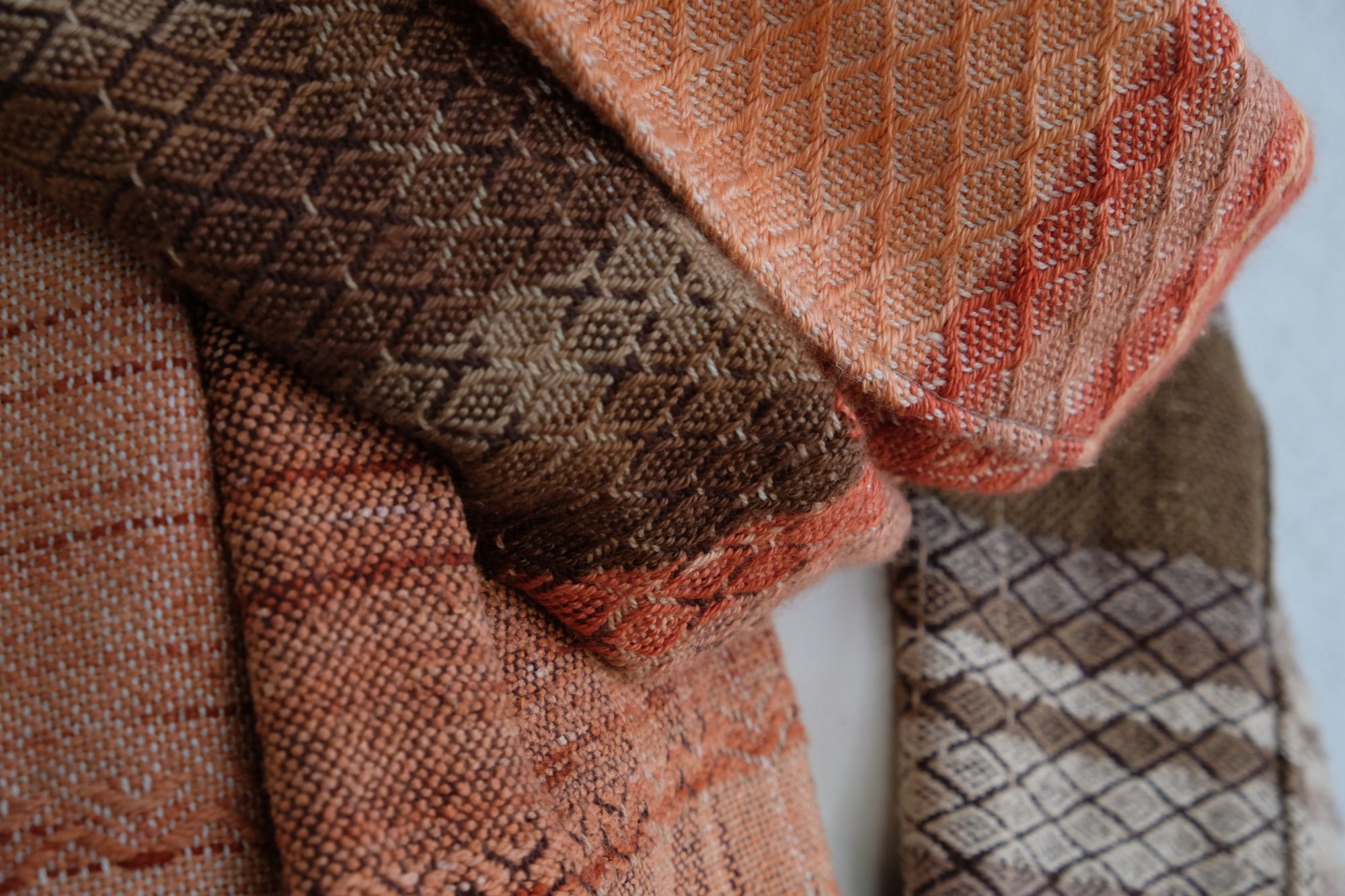 A handwoven shawl is draped on a white painted mannequin against a white wall. The shawl is naturally dyed in shades of earthy browns, oranges and peach pinks