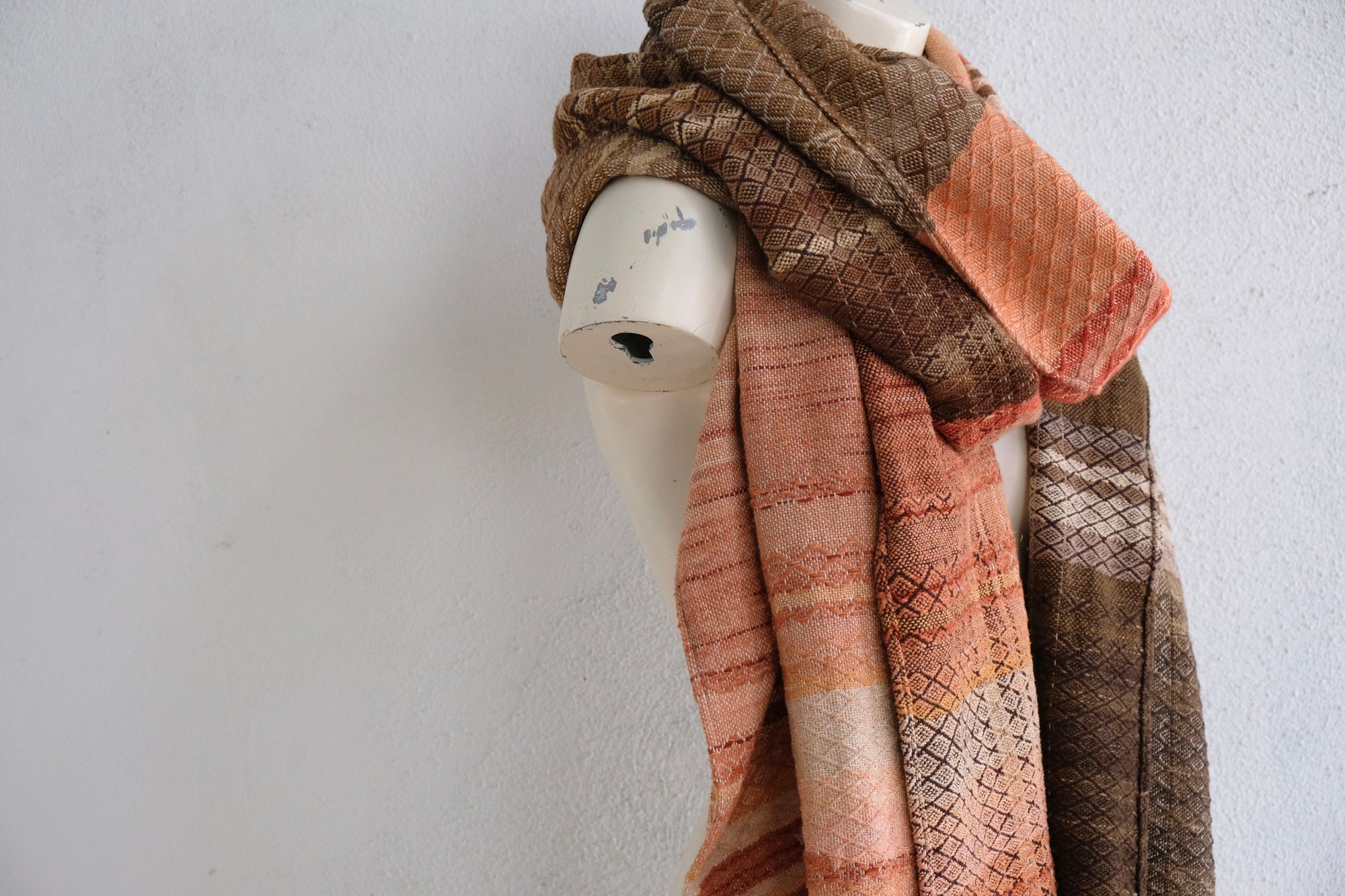 A handwoven shawl is draped on a white painted mannequin against a white wall. The shawl is naturally dyed in shades of earthy browns, oranges and peach pinks