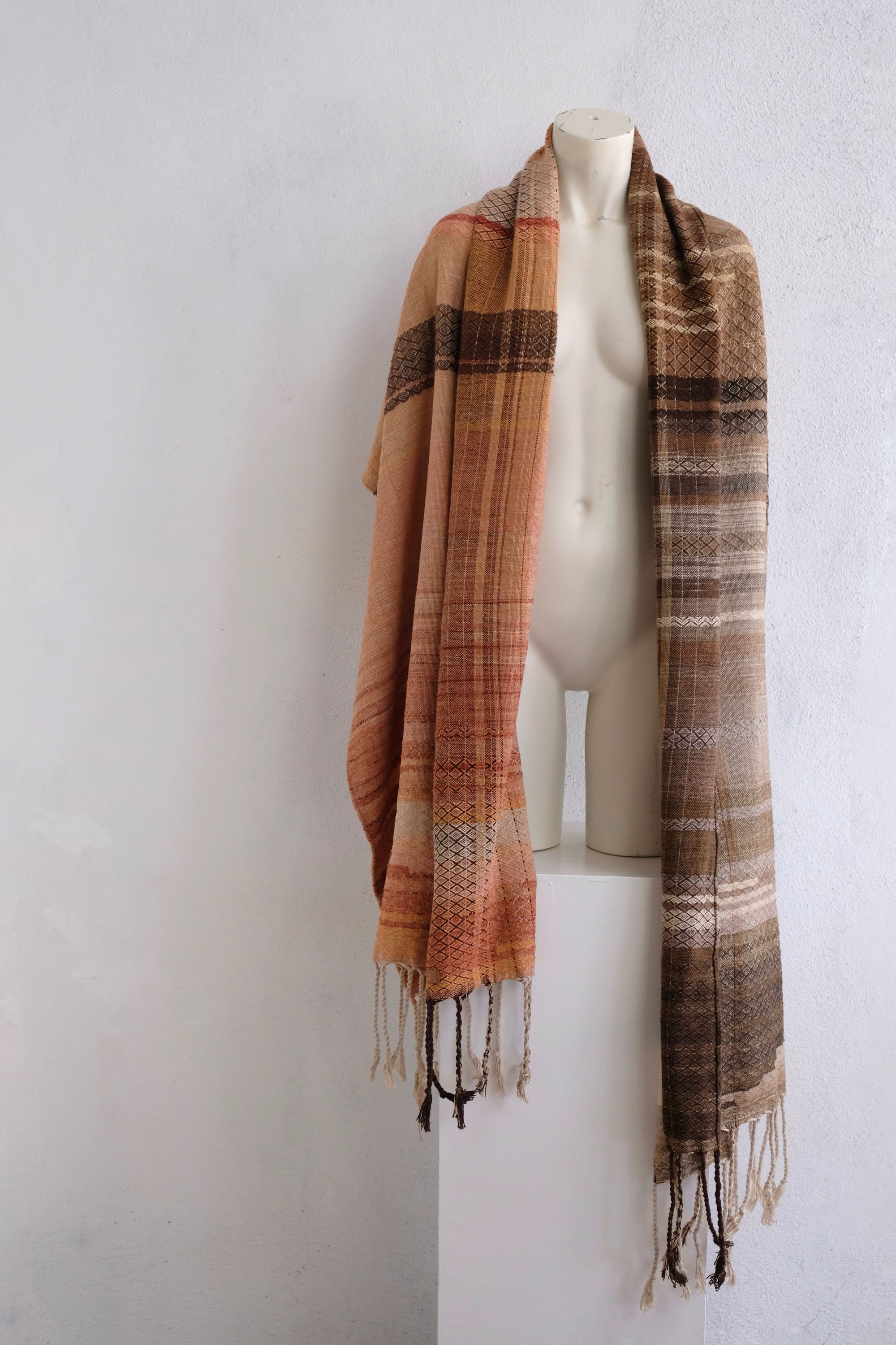 A handwoven shawl is draped on a white painted mannequin against a white wall. The shawl is naturally dyed in shades of earthy browns, oranges and peach pinks