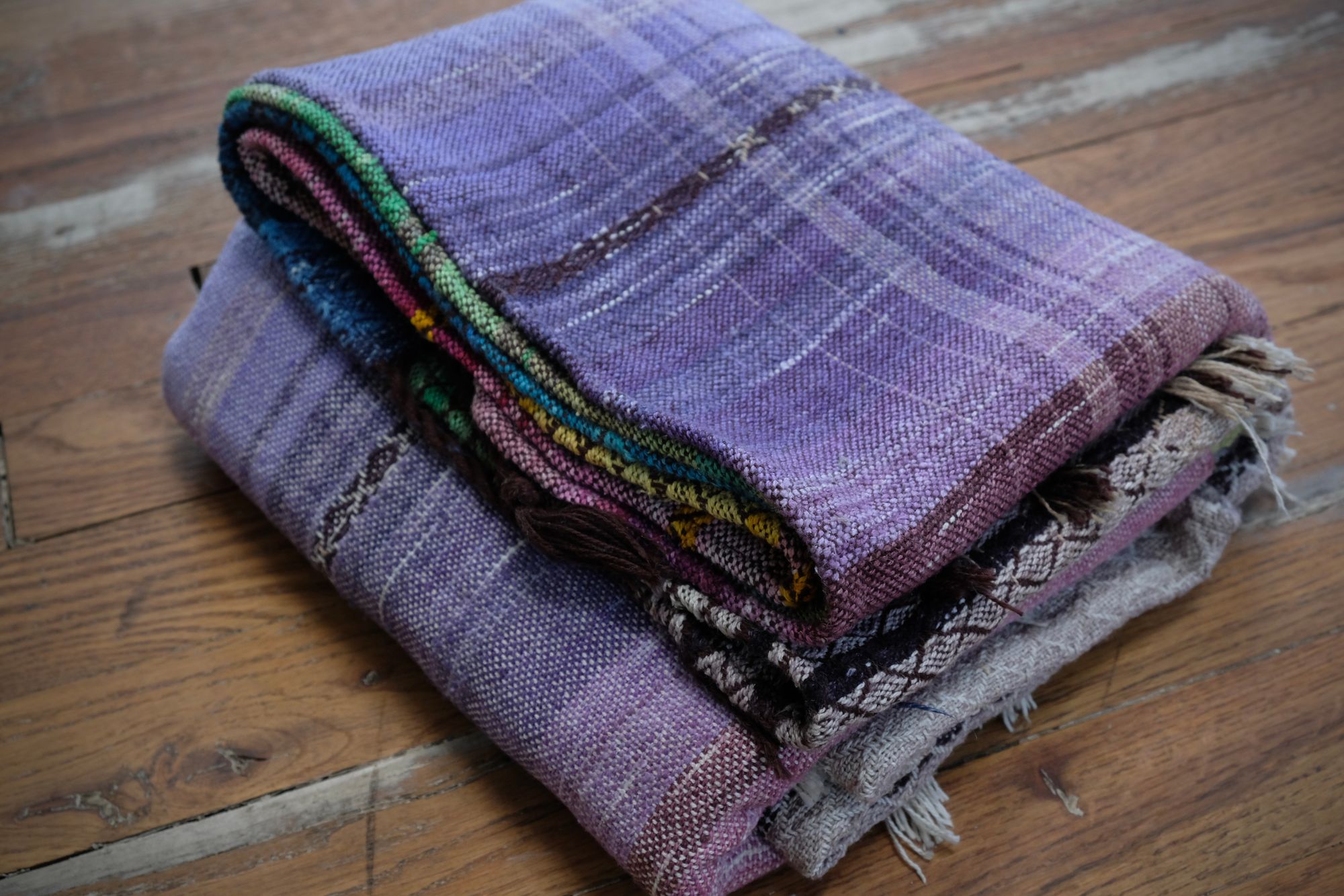 handwoven, hand dyed fabric of many colors with a diamond pattern, folded on a wooden floor. Here the color you see is purple