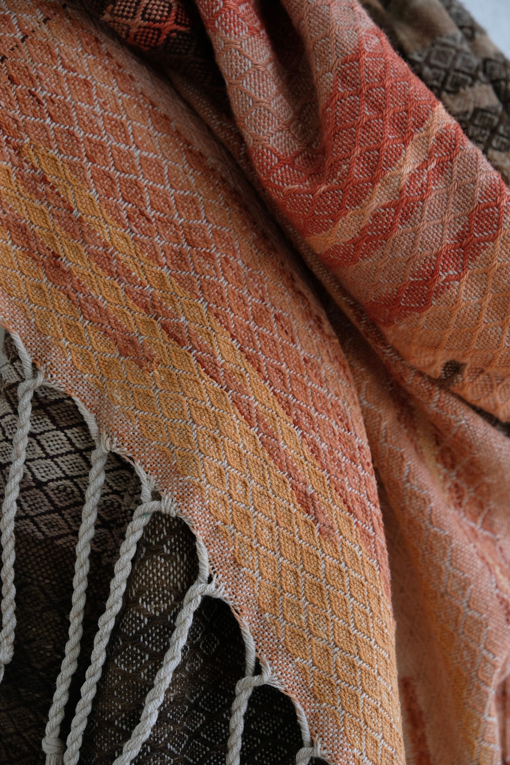 A handwoven shawl is draped on a white painted mannequin against a white wall. The shawl is naturally dyed in shades of earthy browns, oranges and peach pinks