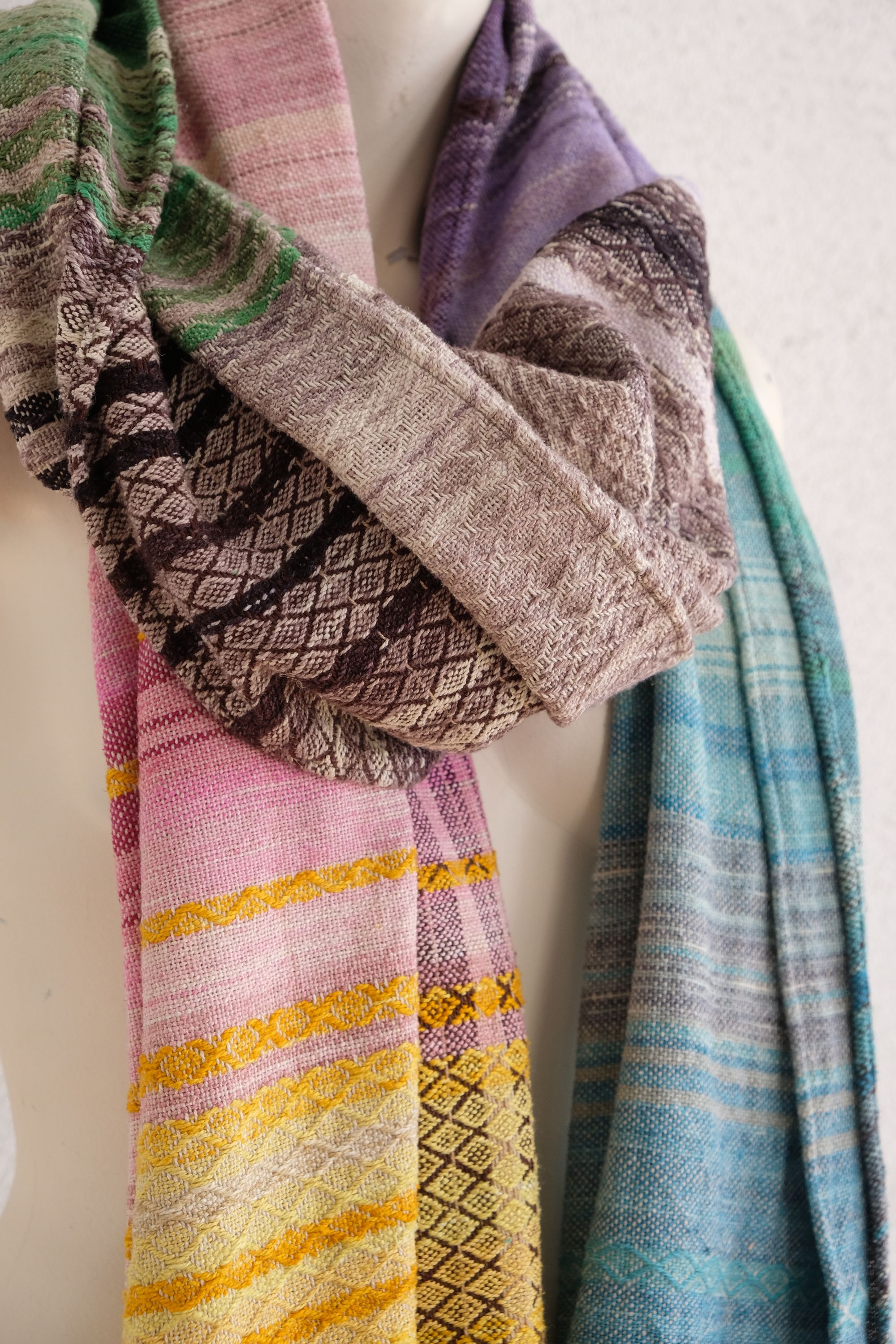 handwoven, hand dyed in blues, green, gray, black, purple, pink and yellow,  fabric of many colors with a diamond pattern, is draped on a mannequin in a white gallery space