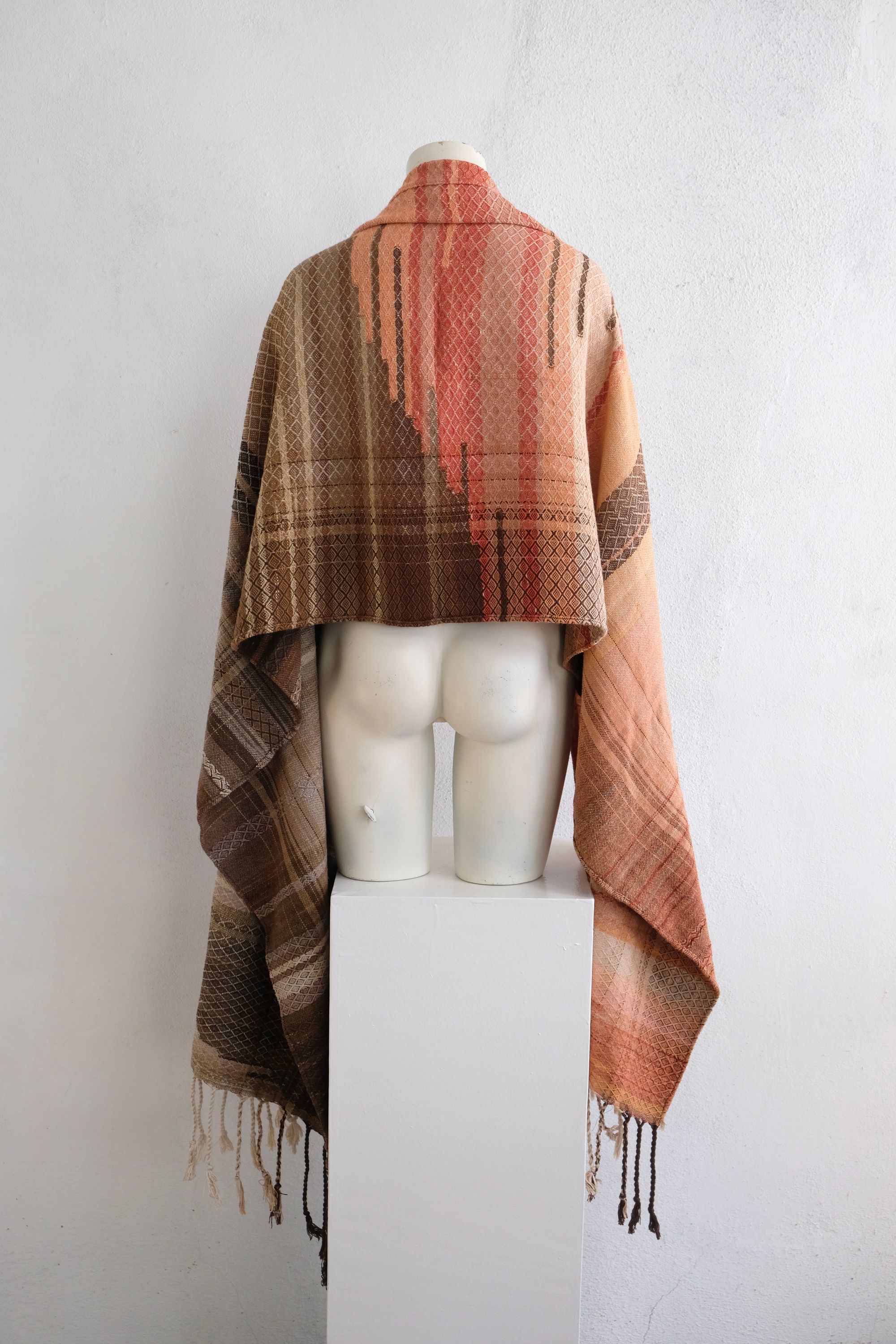 A handwoven shawl is draped on a white painted mannequin against a white wall. The shawl is naturally dyed in shades of earthy browns, oranges and peach pinks
