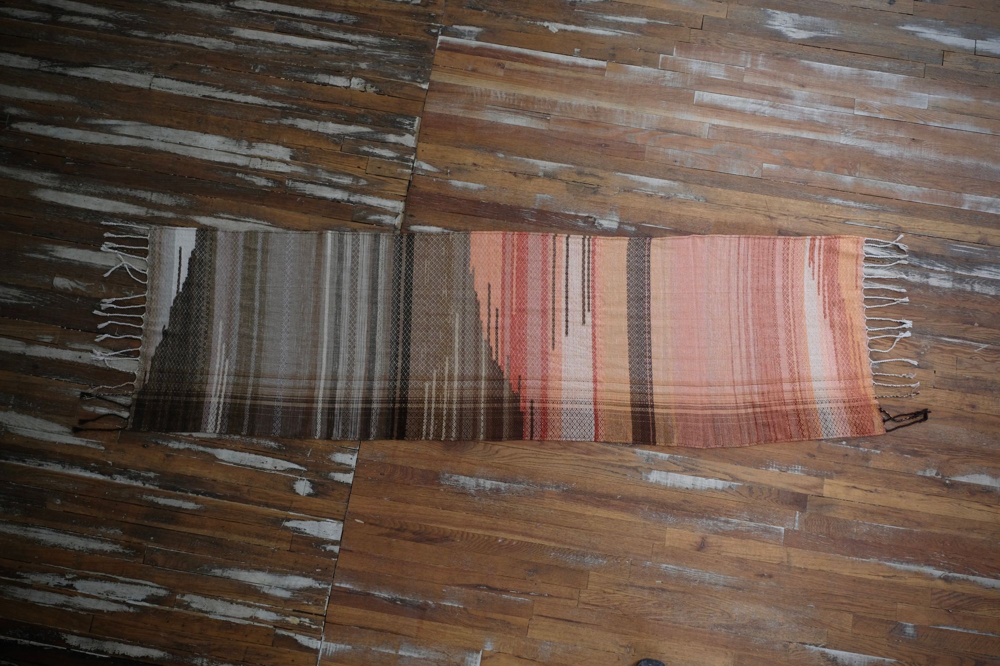 A handwoven shawl is draped on a white painted mannequin against a white wall. The shawl is naturally dyed in shades of earthy browns, oranges and peach pinks