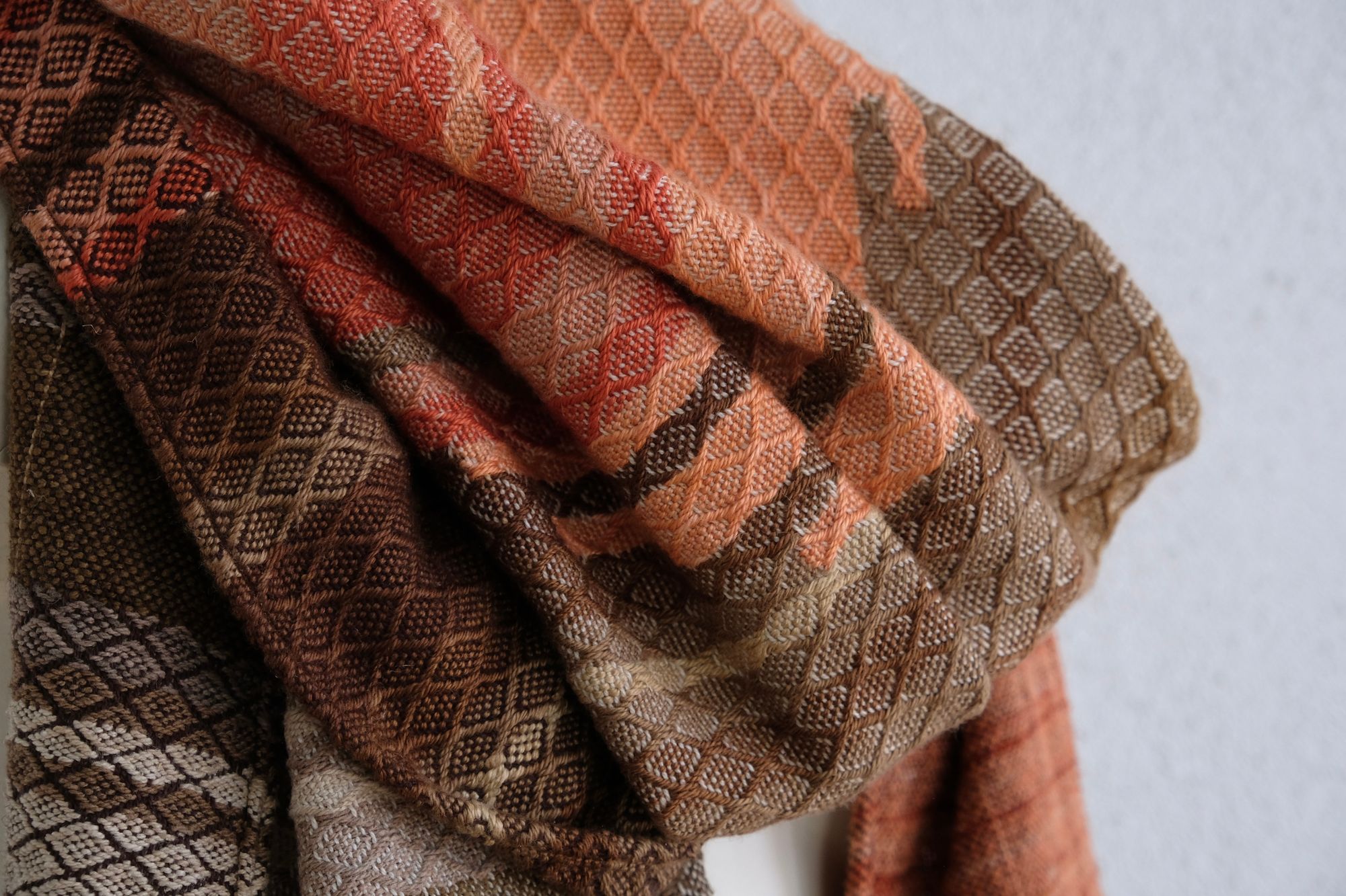 A handwoven shawl is draped on a white painted mannequin against a white wall. The shawl is naturally dyed in shades of earthy browns, oranges and peach pinks