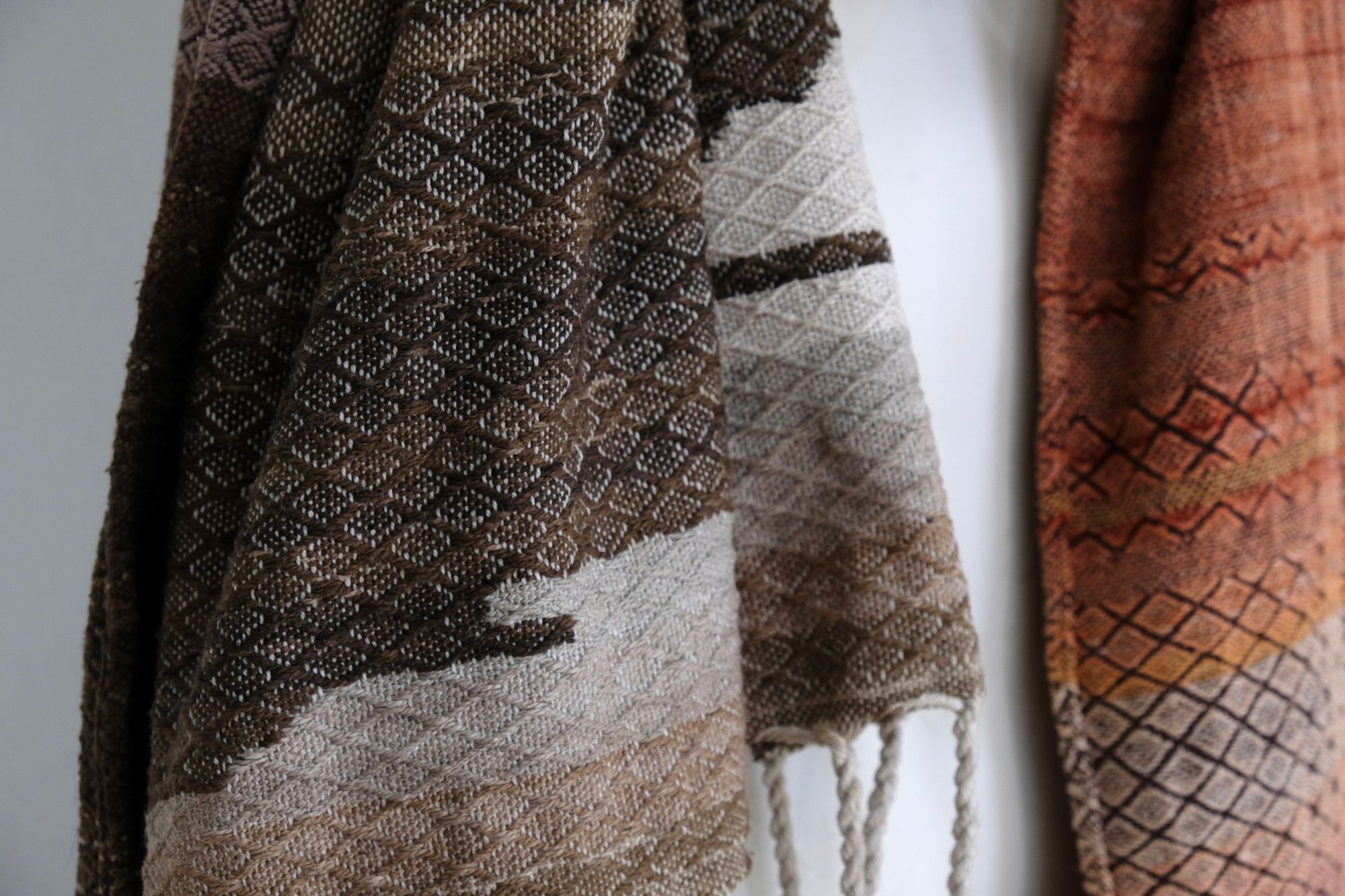 A handwoven shawl is draped on a white painted mannequin against a white wall. The shawl is naturally dyed in shades of earthy browns, oranges and peach pinks
