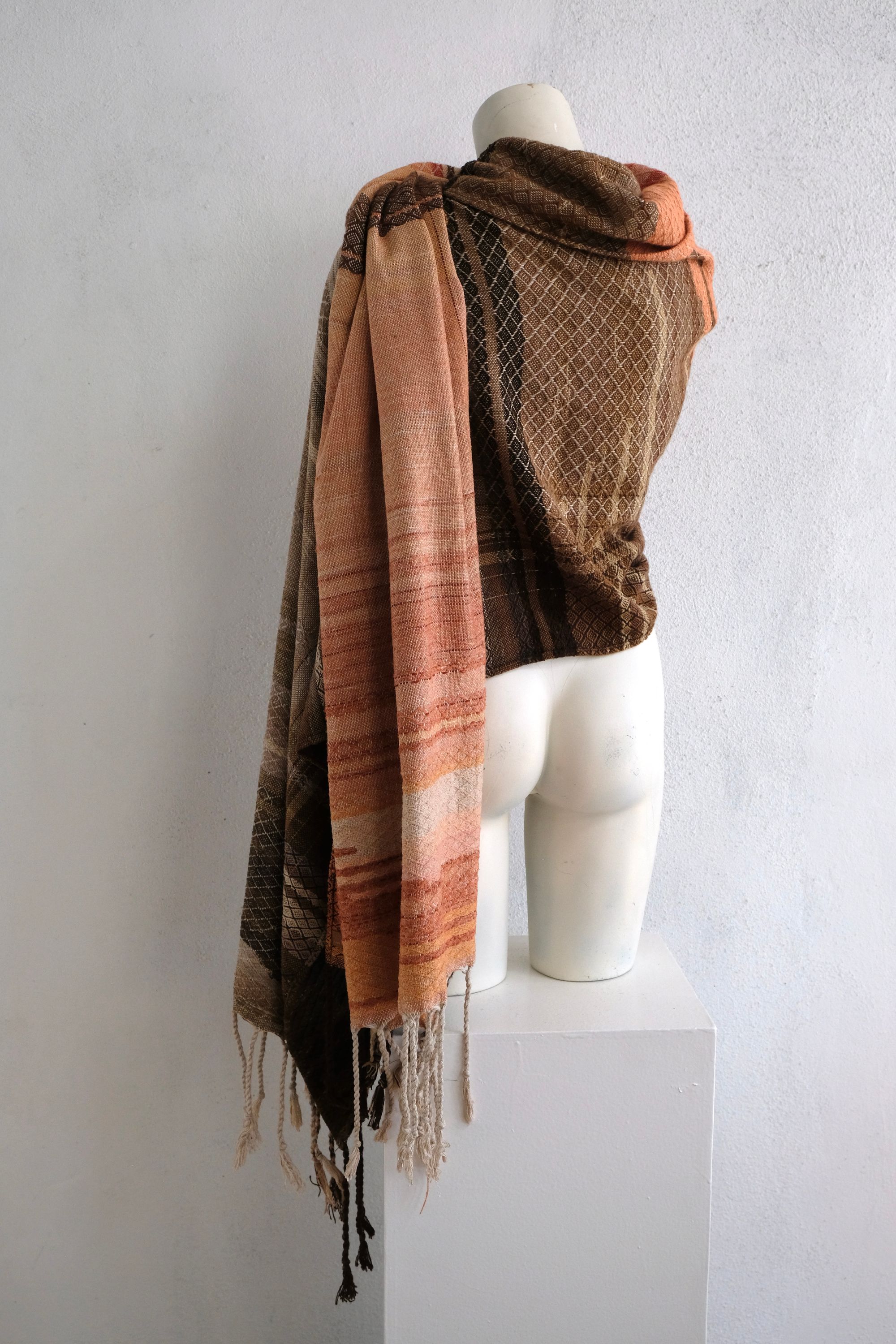 A handwoven shawl is draped on a white painted mannequin against a white wall. The shawl is naturally dyed in shades of earthy browns, oranges and peach pinks