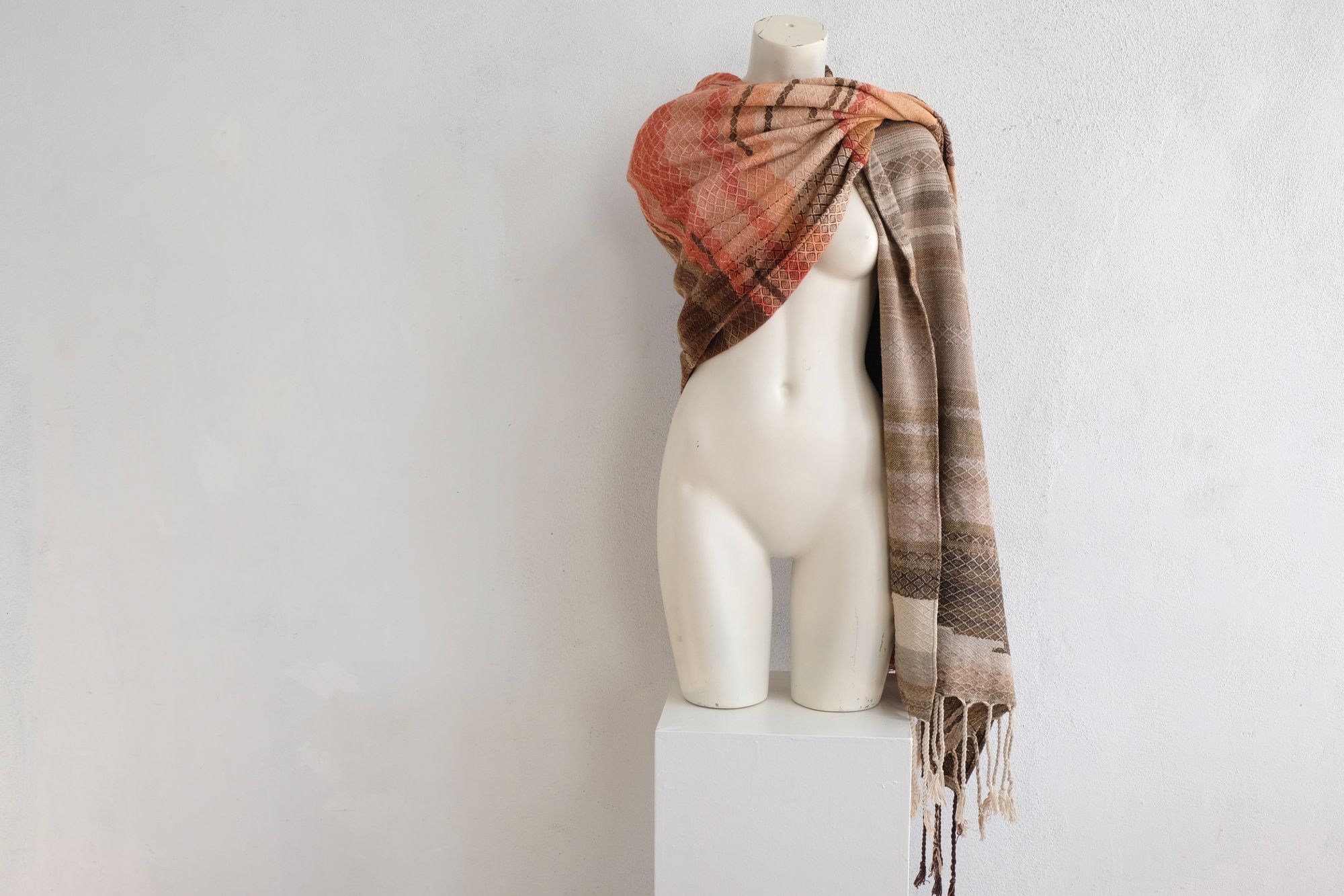 A handwoven shawl is draped on a white painted mannequin against a white wall. The shawl is naturally dyed in shades of earthy browns, oranges and peach pinks
