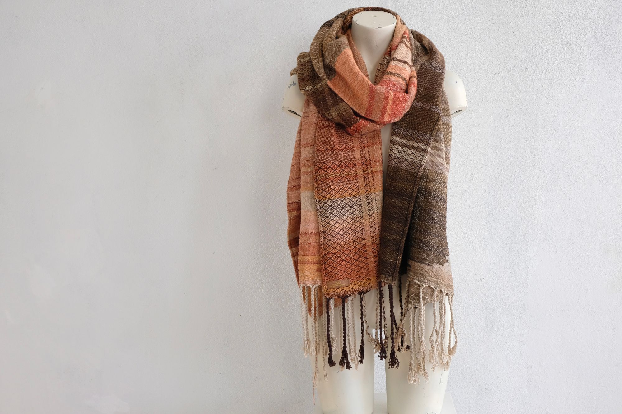 A handwoven shawl is draped on a white painted mannequin against a white wall. The shawl is naturally dyed in shades of earthy browns, oranges and peach pinks