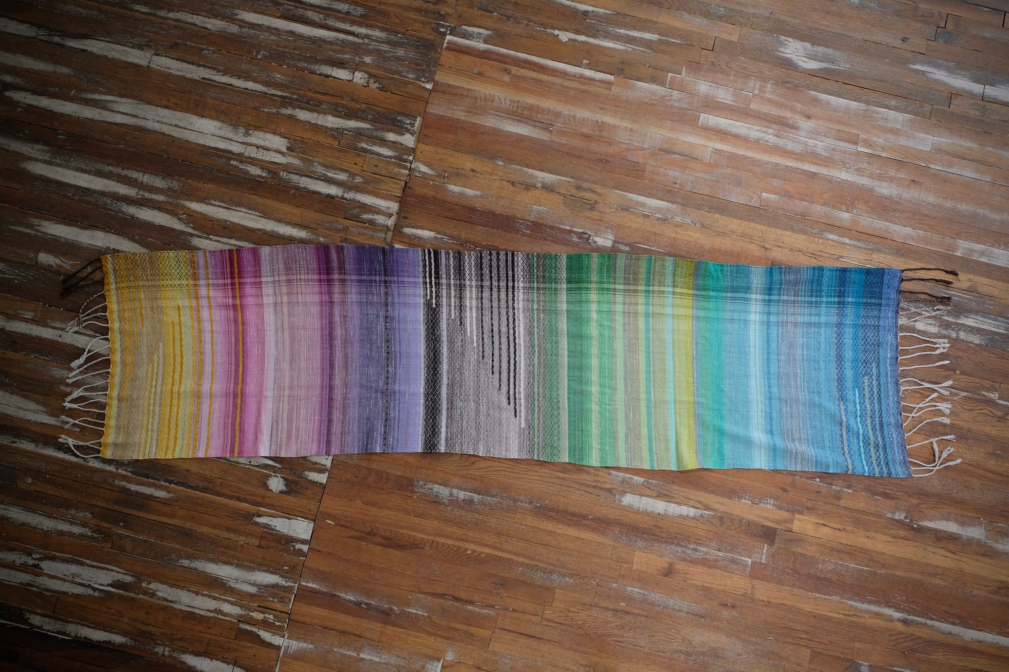 handwoven, hand dyed in blues, green, gray, black, purple, pink and yellow,  fabric of many colors with a diamond pattern, laid out on a wooden floor.