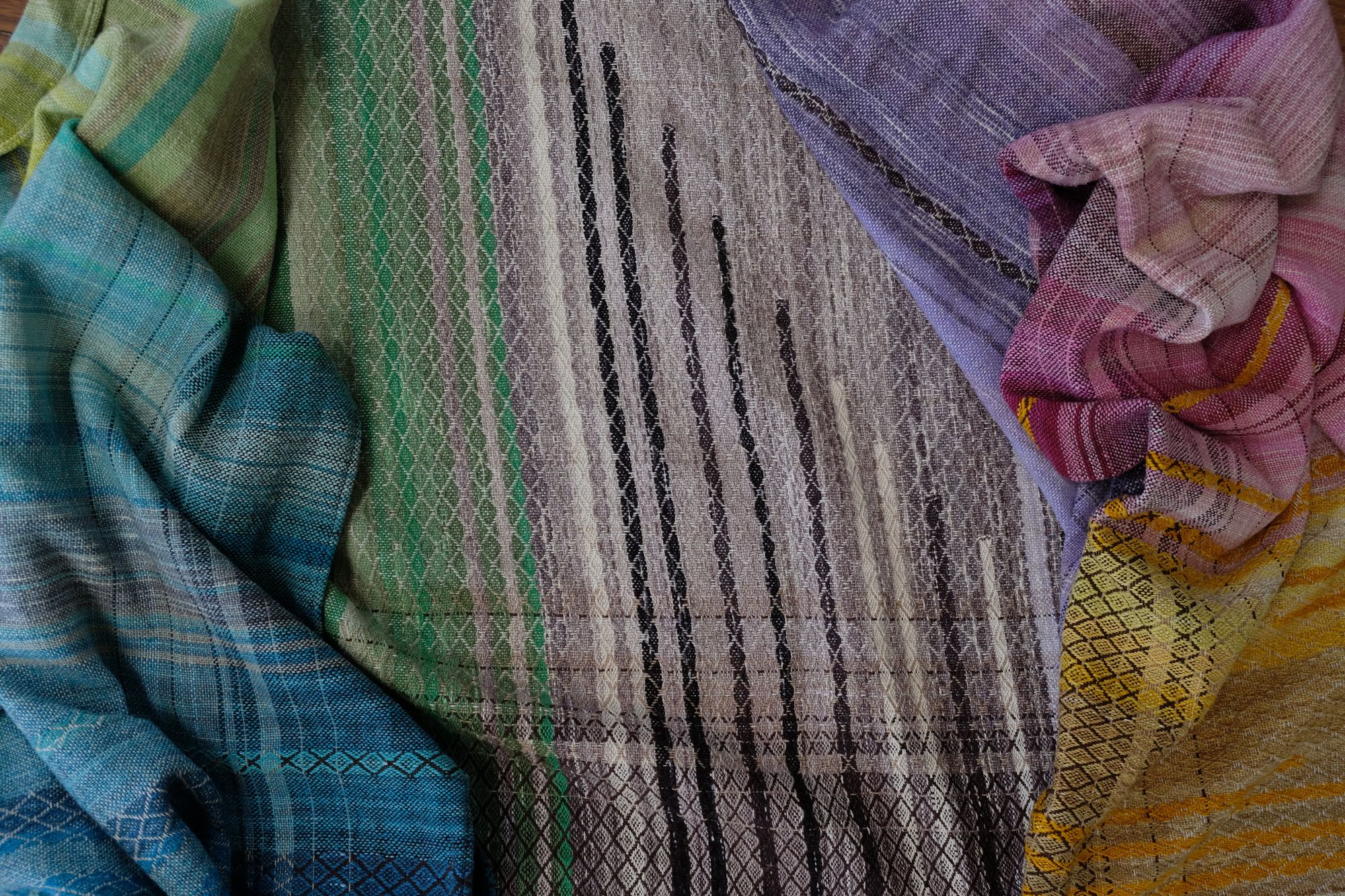 handwoven, hand dyed in blues, green, gray, black, purple, pink and yellow,  fabric of many colors with a diamond pattern, laid out on a wooden floor.