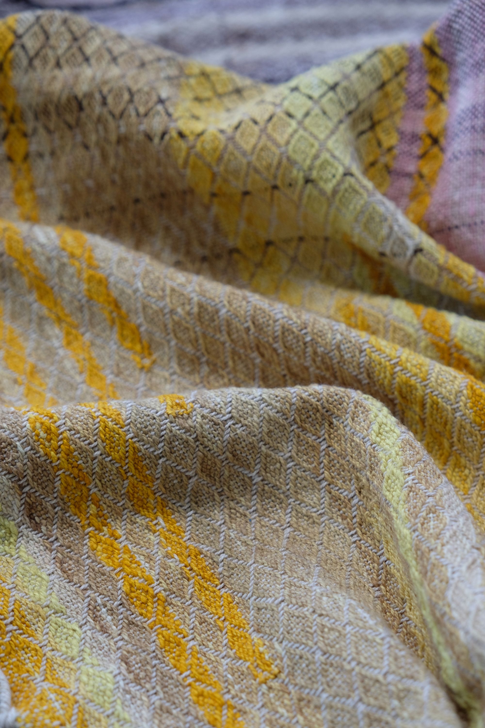 handwoven, hand dyed fabric of many colors with a diamond pattern, laid out on a wooden floor.